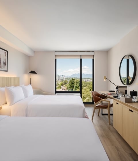 Hotel room with two queen beds, beige headboard, desk, and a round wall mirror overlooking a large window with city and mountain view.