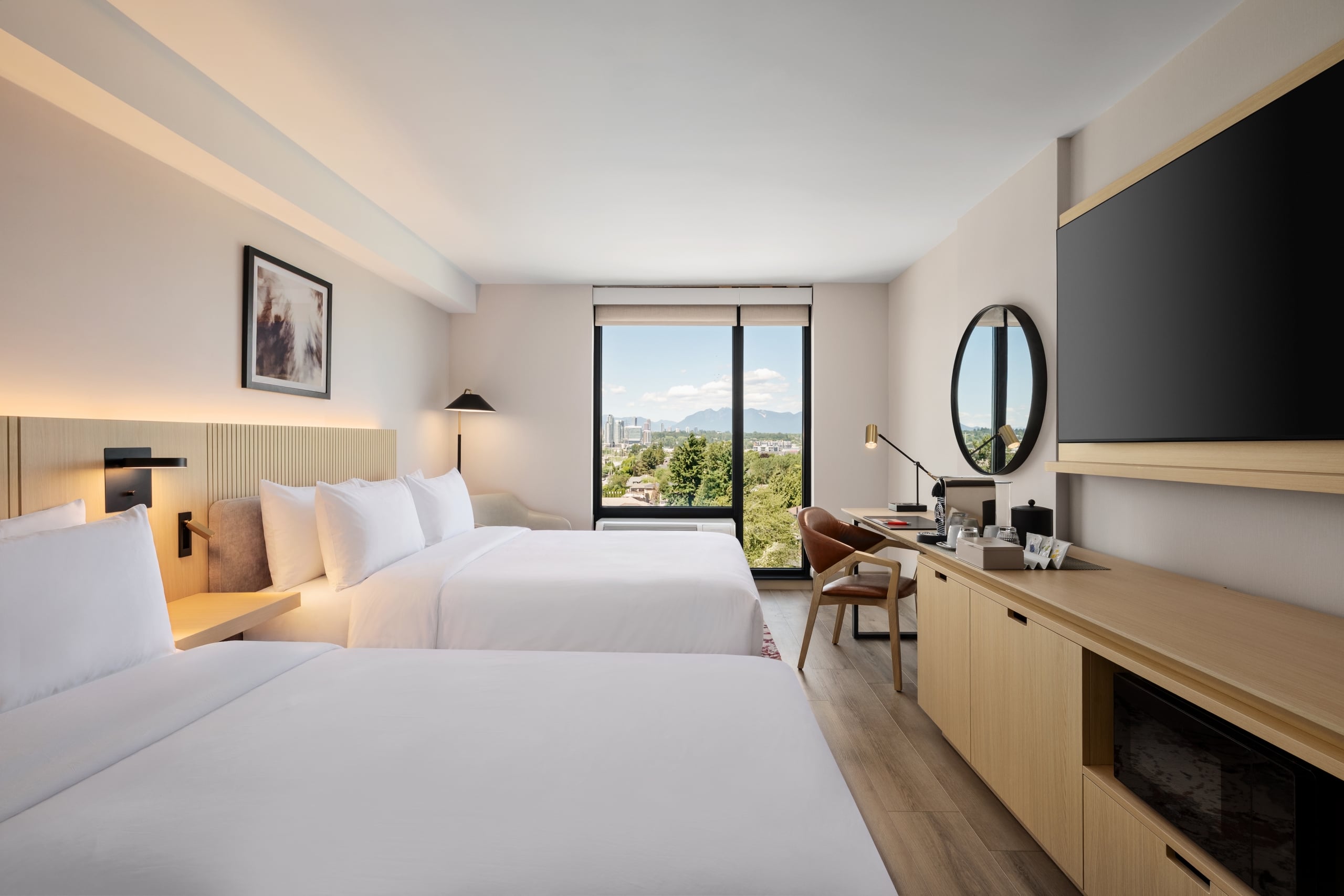 Hotel room with two queen beds, beige headboard, desk, and a round wall mirror overlooking a large window with city and mountain view.