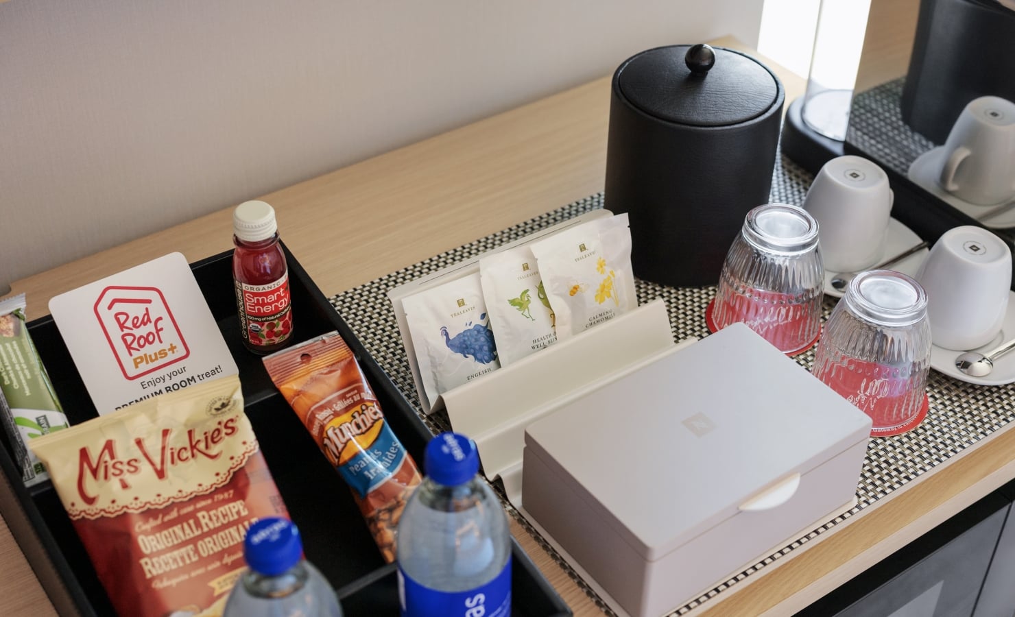Hotel minibar with bottled water, assorted snacks, and tea sachets arranged on a tray, plus a black electric kettle nearby.