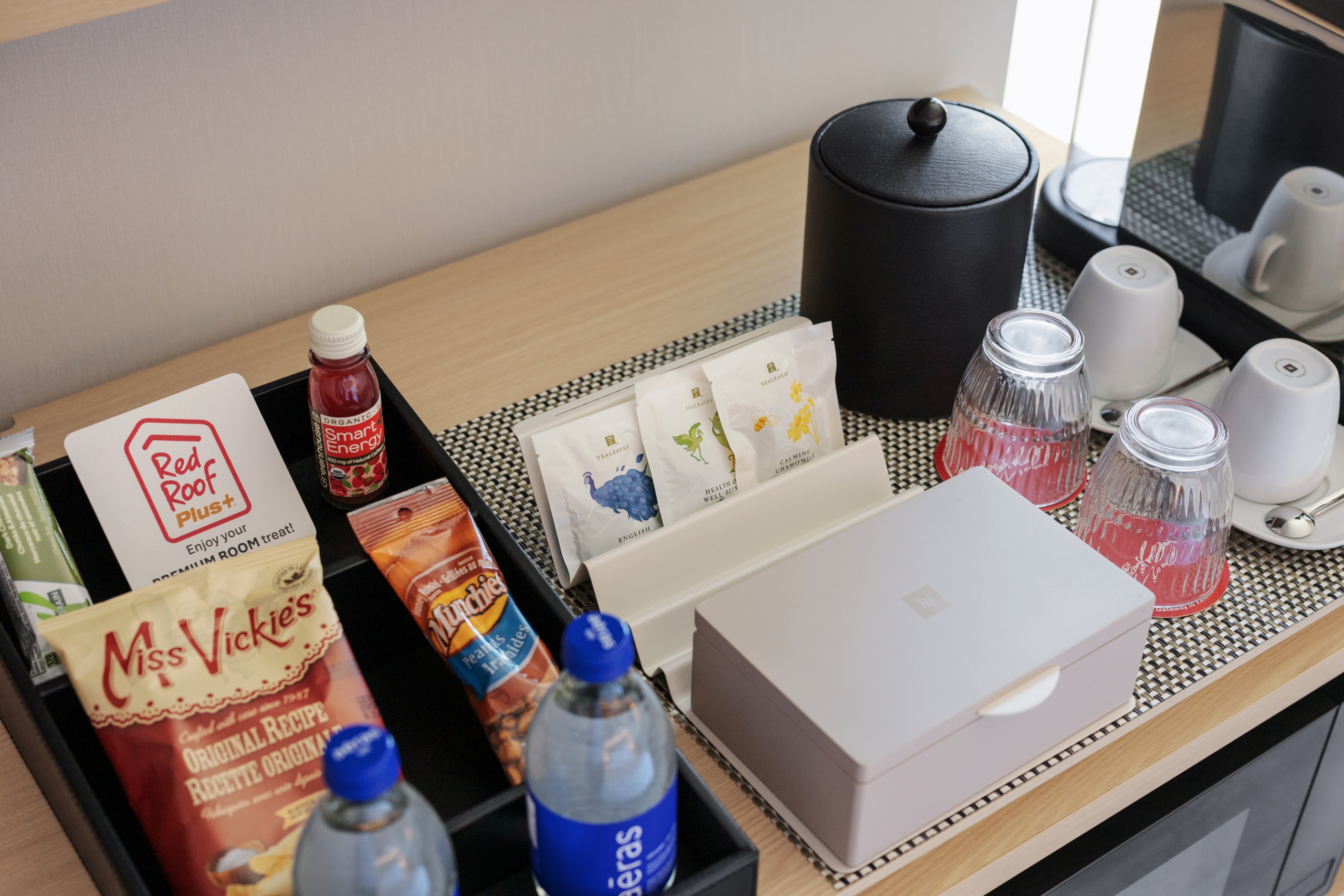Hotel minibar with bottled water, assorted snacks, and tea sachets arranged on a tray, plus a black electric kettle nearby.