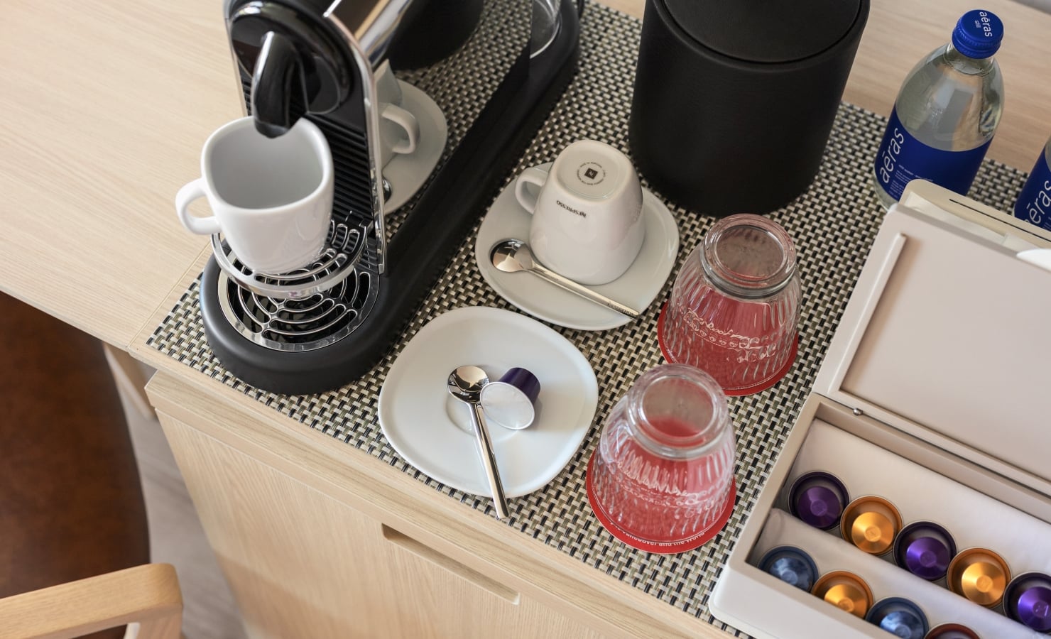 Open kitchen drawer showing a coffee station: espresso machine, white mug on saucer, spoons, glass jars, and colorful coffee capsules in trays and a bottle nearby.