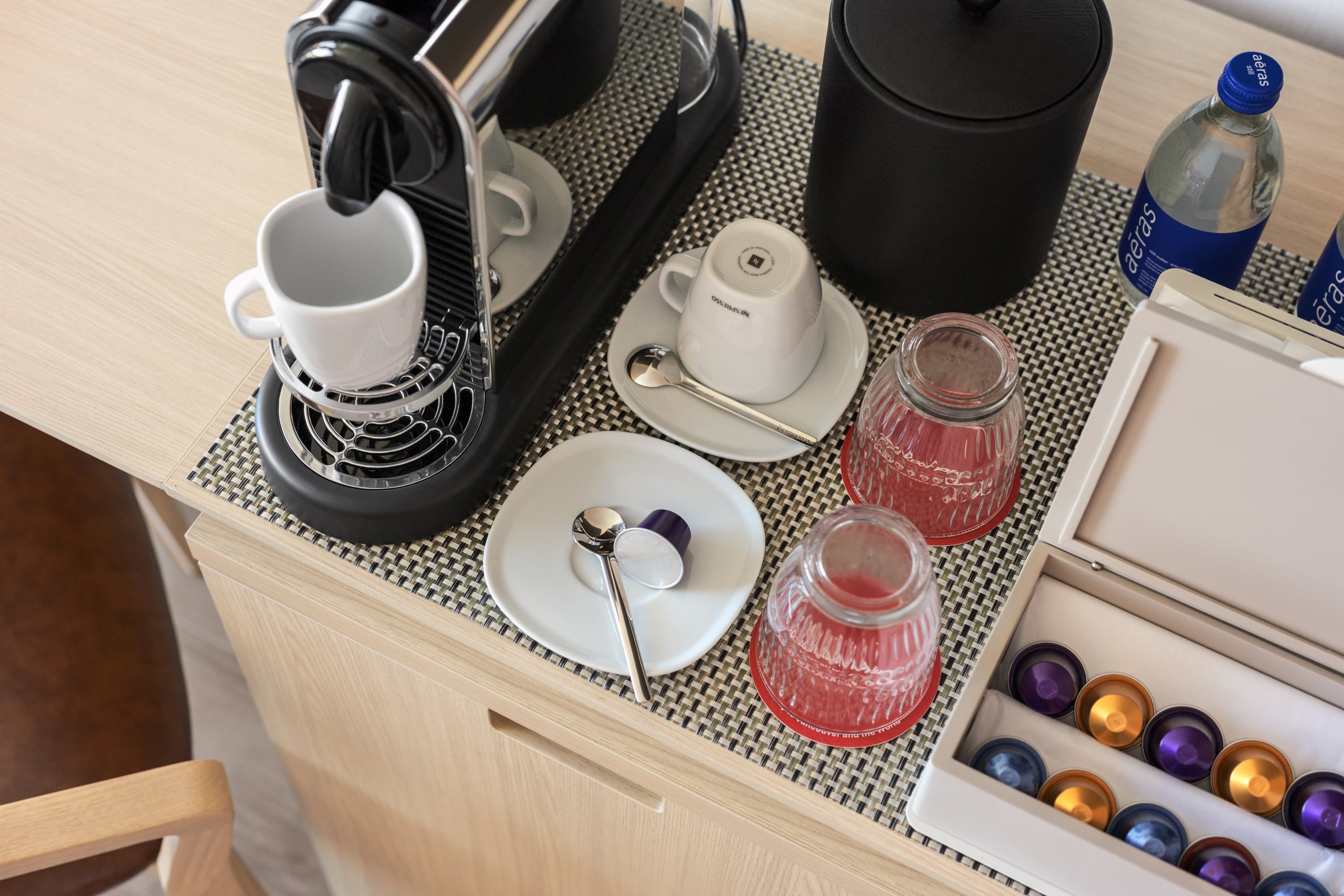 Open kitchen drawer showing a coffee station: espresso machine, white mug on saucer, spoons, glass jars, and colorful coffee capsules in trays and a bottle nearby.