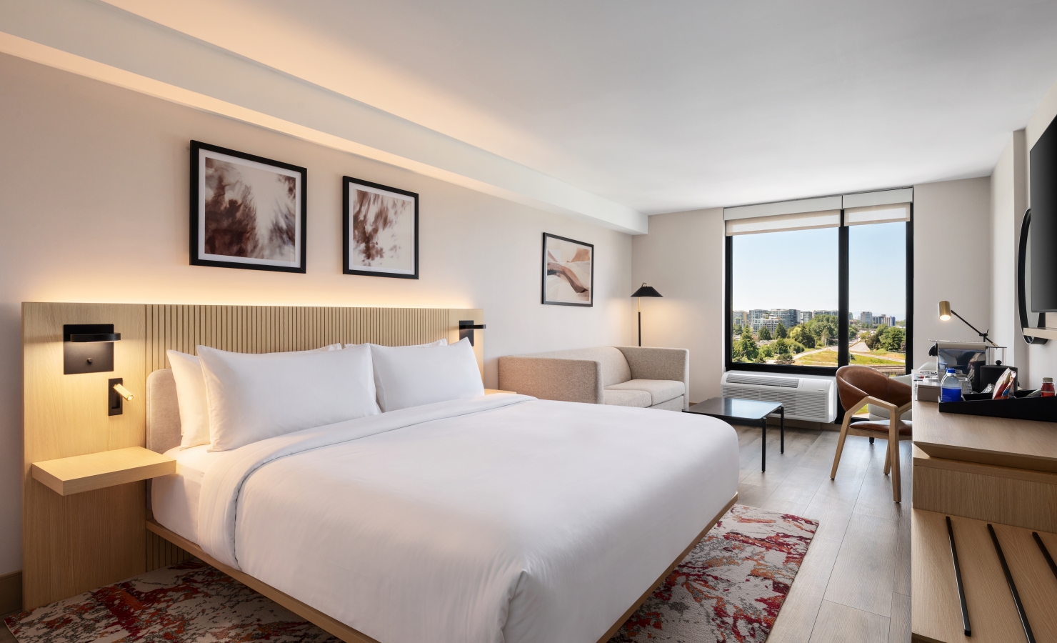 Modern hotel room with a large white bed, wooden headboard, and bedside lamps; a seating area and a work desk near a window offering a city view.
