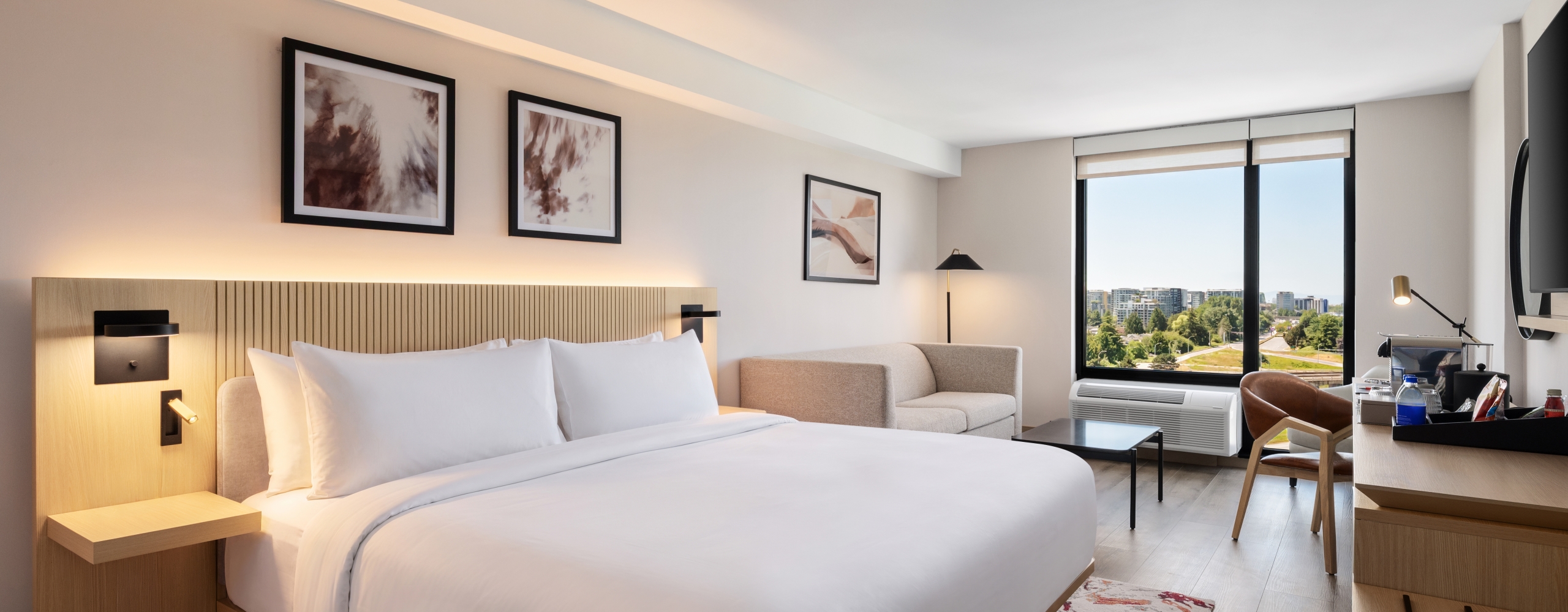 Modern hotel room with a large white bed, wooden headboard, and bedside lamps; a seating area and a work desk near a window offering a city view.