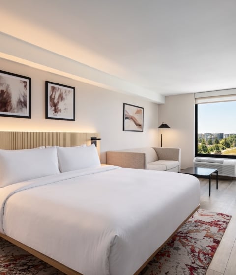 Modern hotel room with a large white bed, wooden headboard, and bedside lamps; a seating area and a work desk near a window offering a city view.