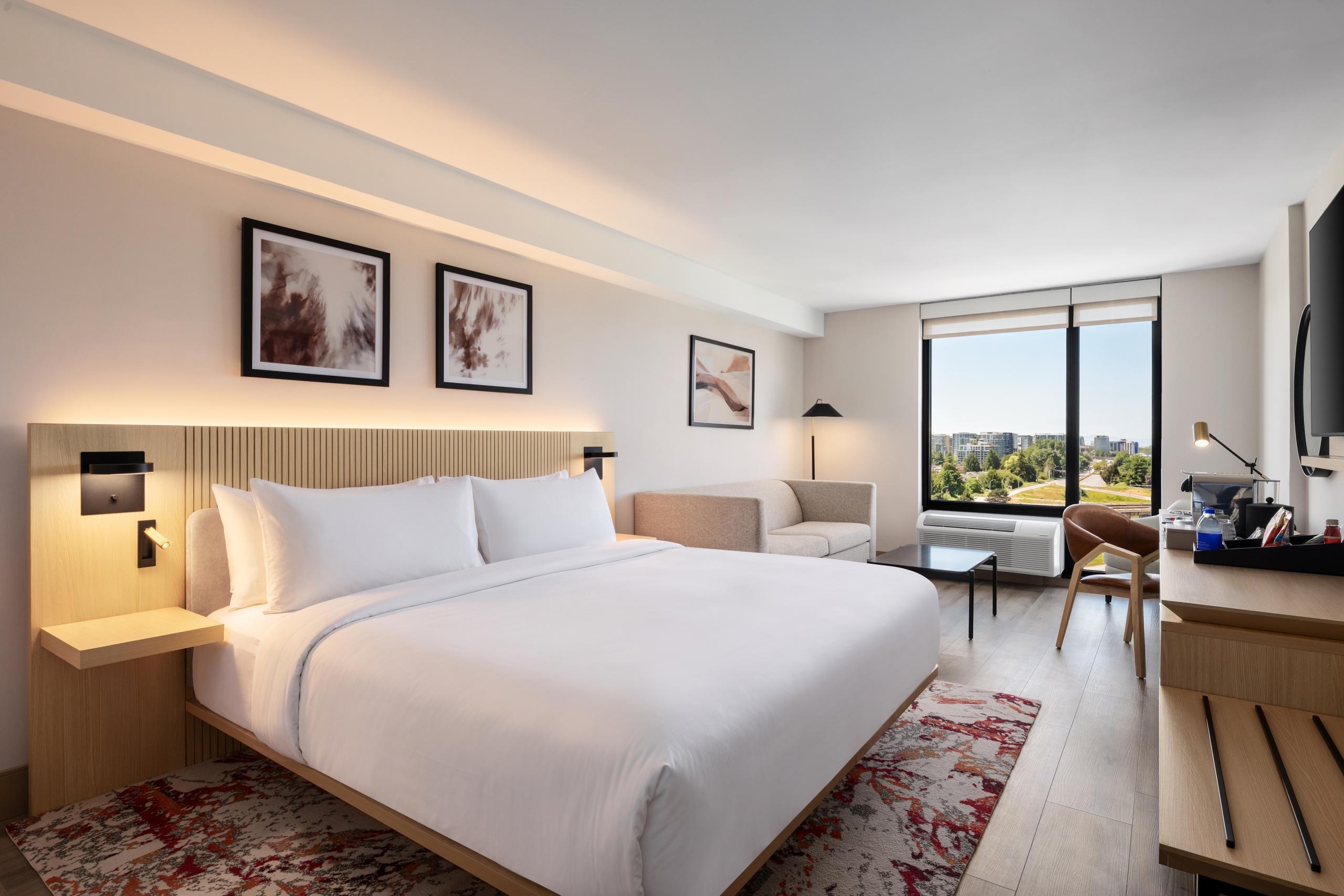 Modern hotel room with a large white bed, wooden headboard, and bedside lamps; a seating area and a work desk near a window offering a city view.