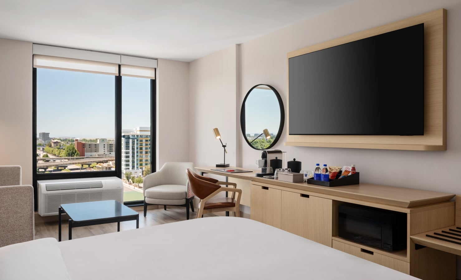 Hotel room with a large flat-screen TV on a wooden mount, overlooking a desk area and city view through tall windows.