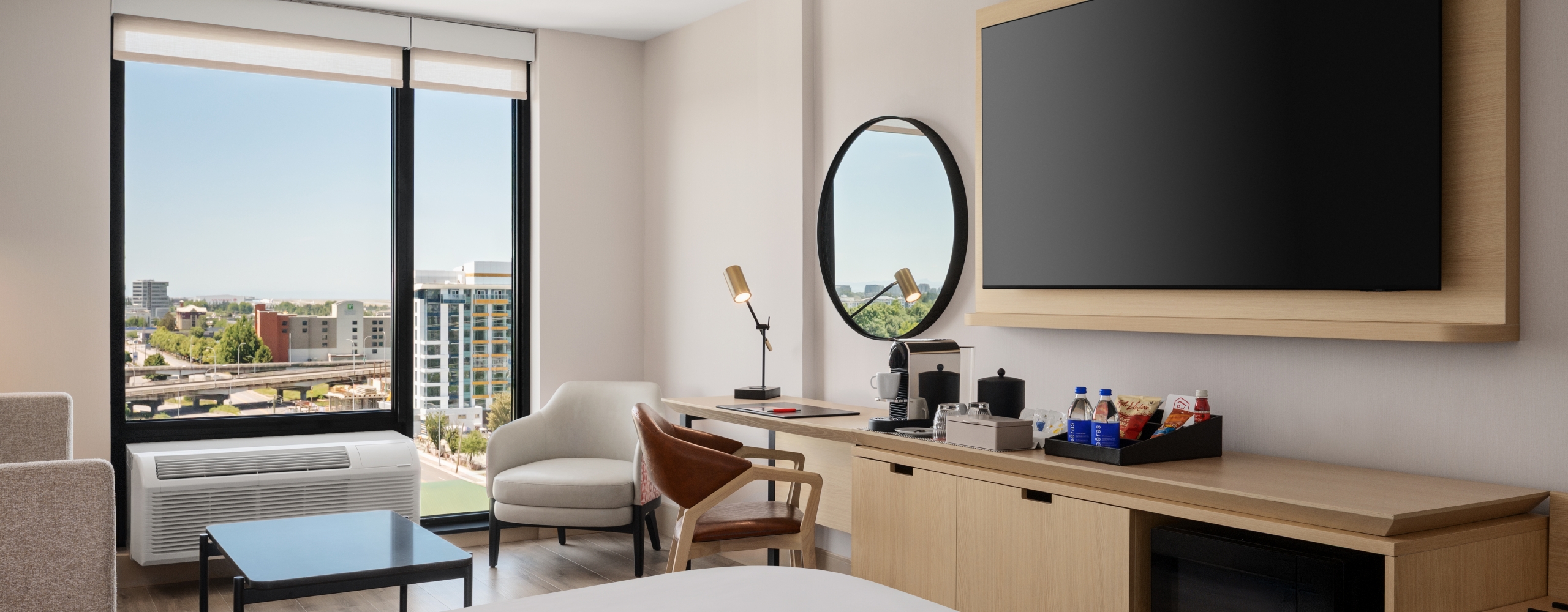 Hotel room with a large flat-screen TV on a wooden mount, overlooking a desk area and city view through tall windows.