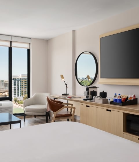 Hotel room with a large flat-screen TV on a wooden mount, overlooking a desk area and city view through tall windows.