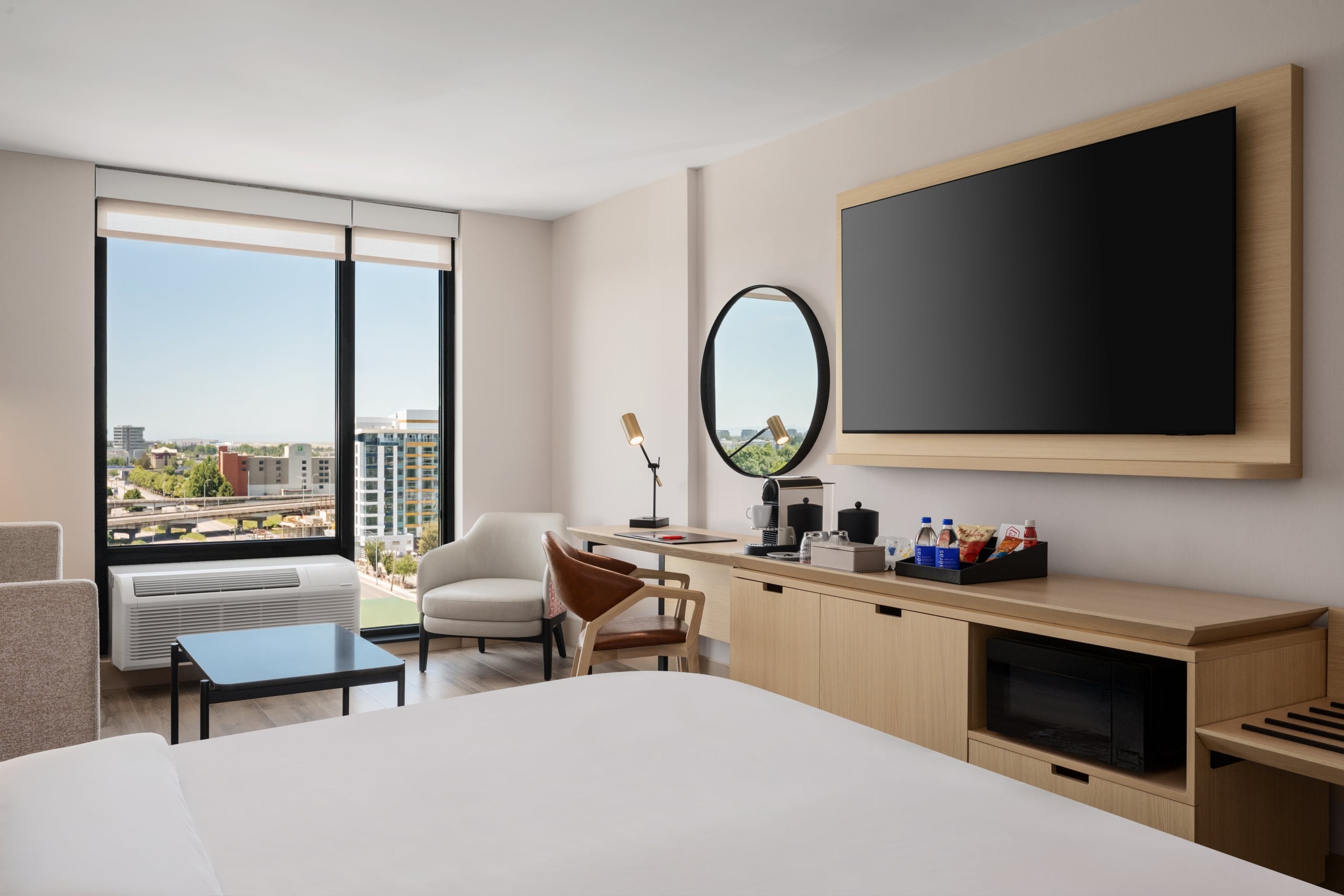 Hotel room with a large flat-screen TV on a wooden mount, overlooking a desk area and city view through tall windows.