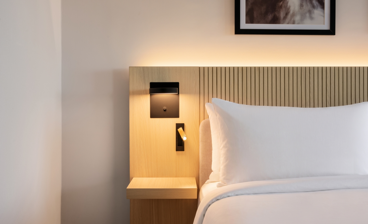 Hotel bed with a vertical slat wood headboard, built-in reading light, and white pillows in a minimalist room.
