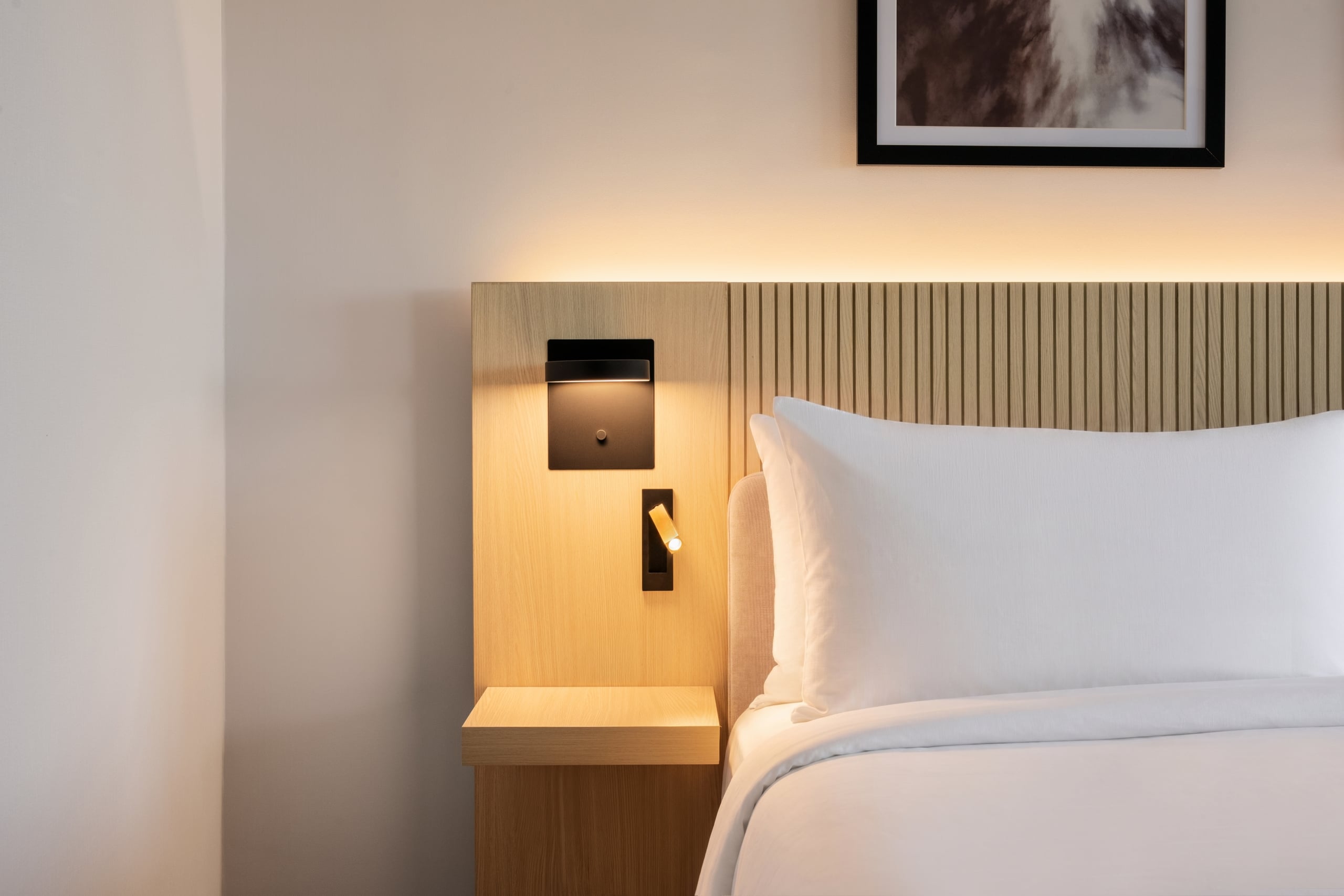 Hotel bed with a vertical slat wood headboard, built-in reading light, and white pillows in a minimalist room.