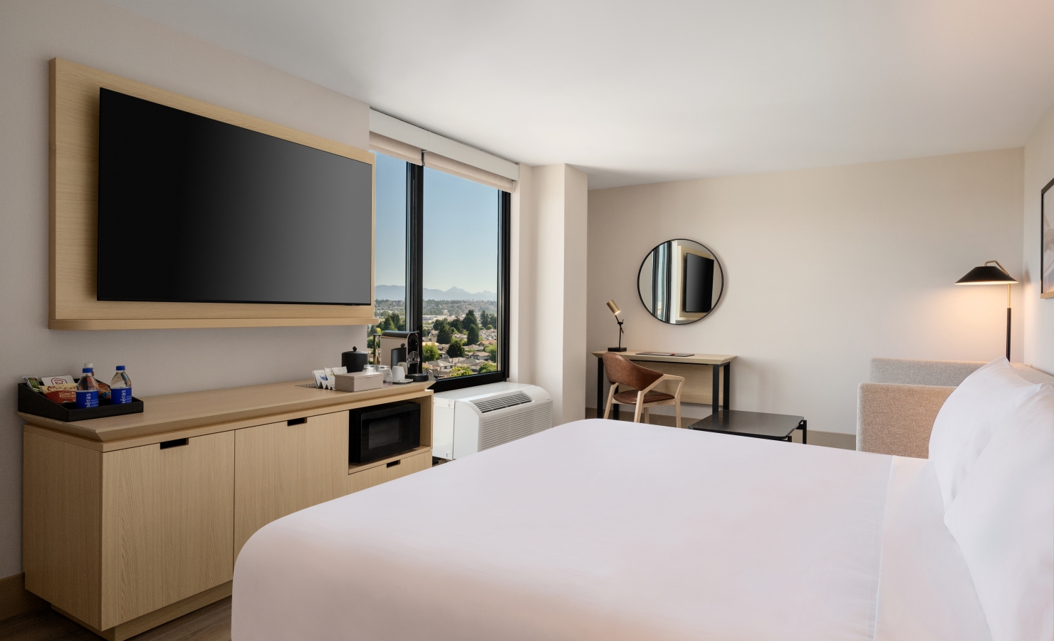 Hotel room with a wall-mounted flat-screen TV on a wooden panel, a desk and mirror near a window with a city and mountain view.