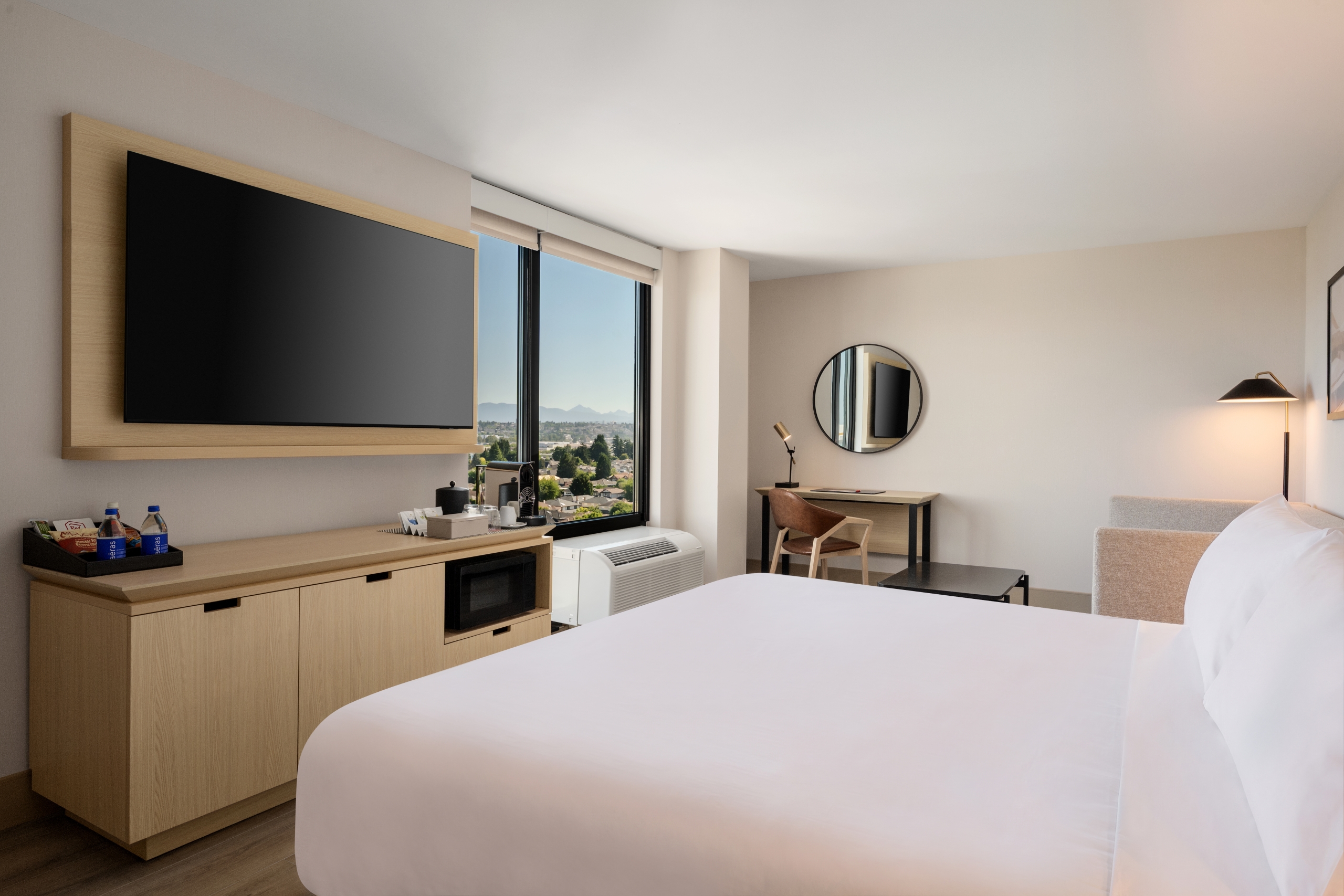 Hotel room with a wall-mounted flat-screen TV on a wooden panel, a desk and mirror near a window with a city and mountain view.