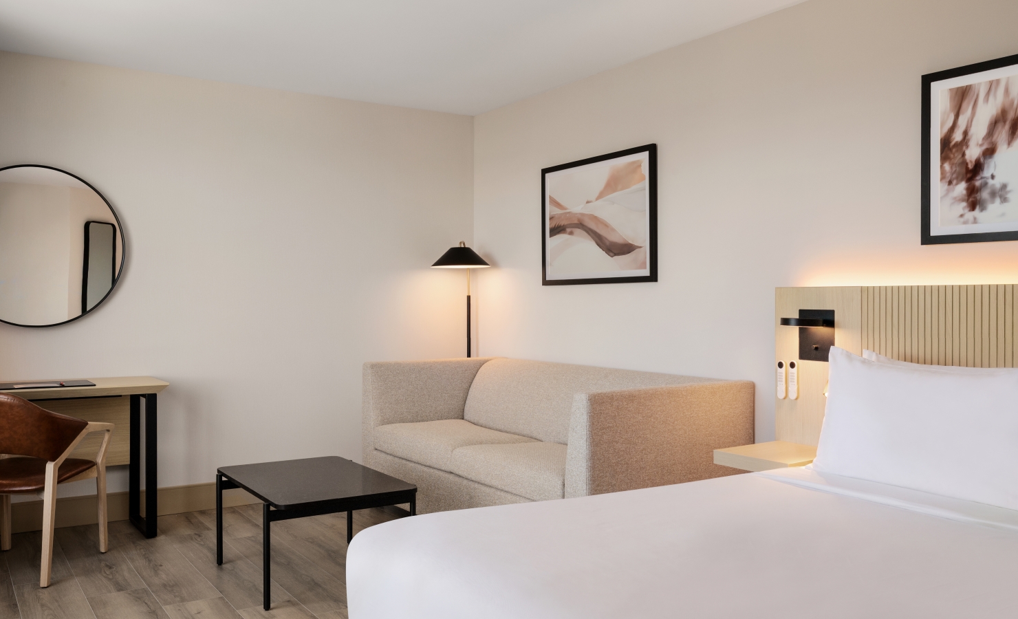 Hotel room interior with a beige sofa in the foreground, round wall mirror on the left, and framed abstract art above the bed area.