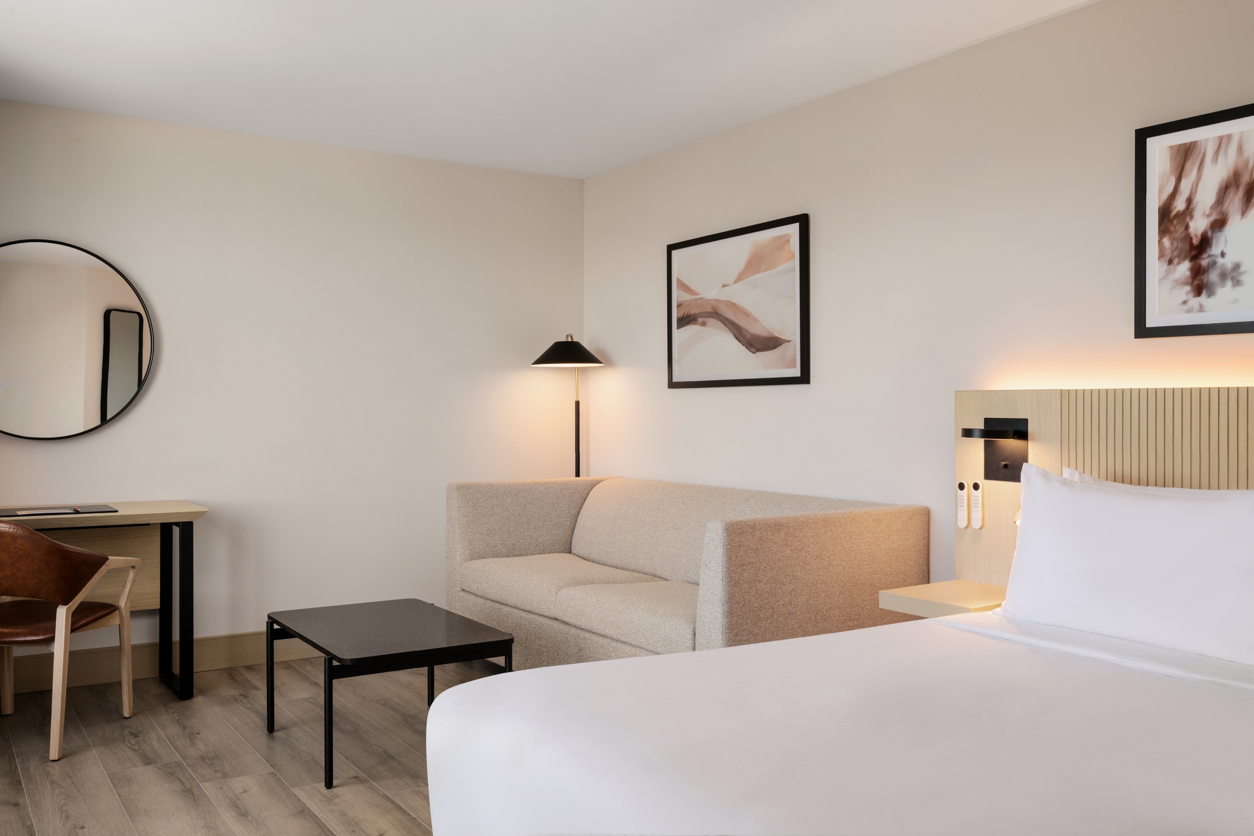Hotel room interior with a beige sofa in the foreground, round wall mirror on the left, and framed abstract art above the bed area.
