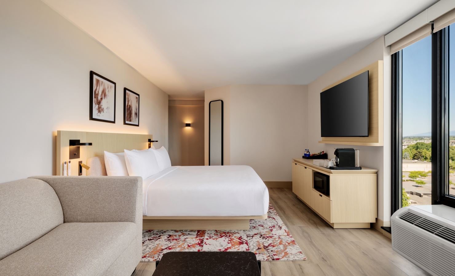 Hotel room with a large bed, beige sofa, wall-mounted TV, and a glass window view of the city outside.