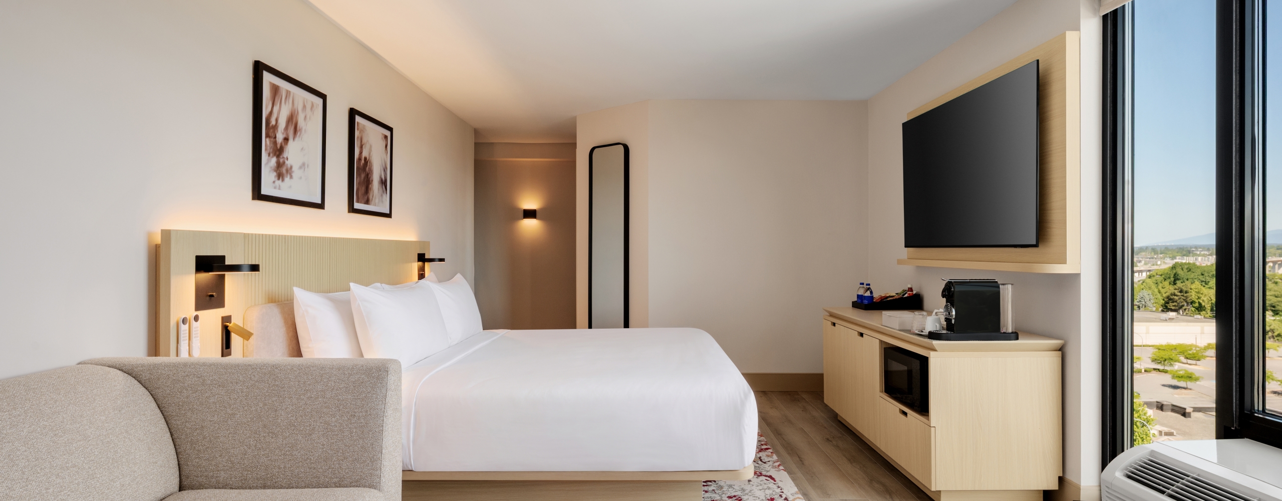 Hotel room with a large bed, beige sofa, wall-mounted TV, and a glass window view of the city outside.