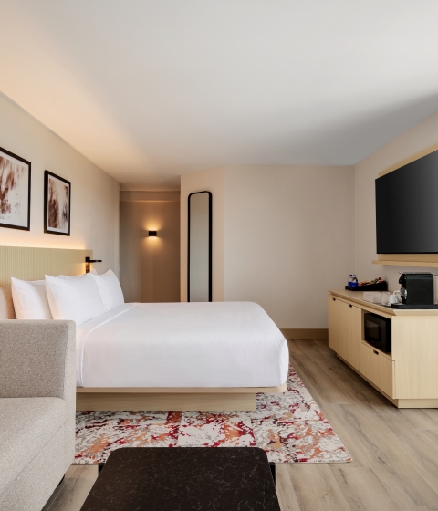 Hotel room with a large bed, beige sofa, wall-mounted TV, and a glass window view of the city outside.