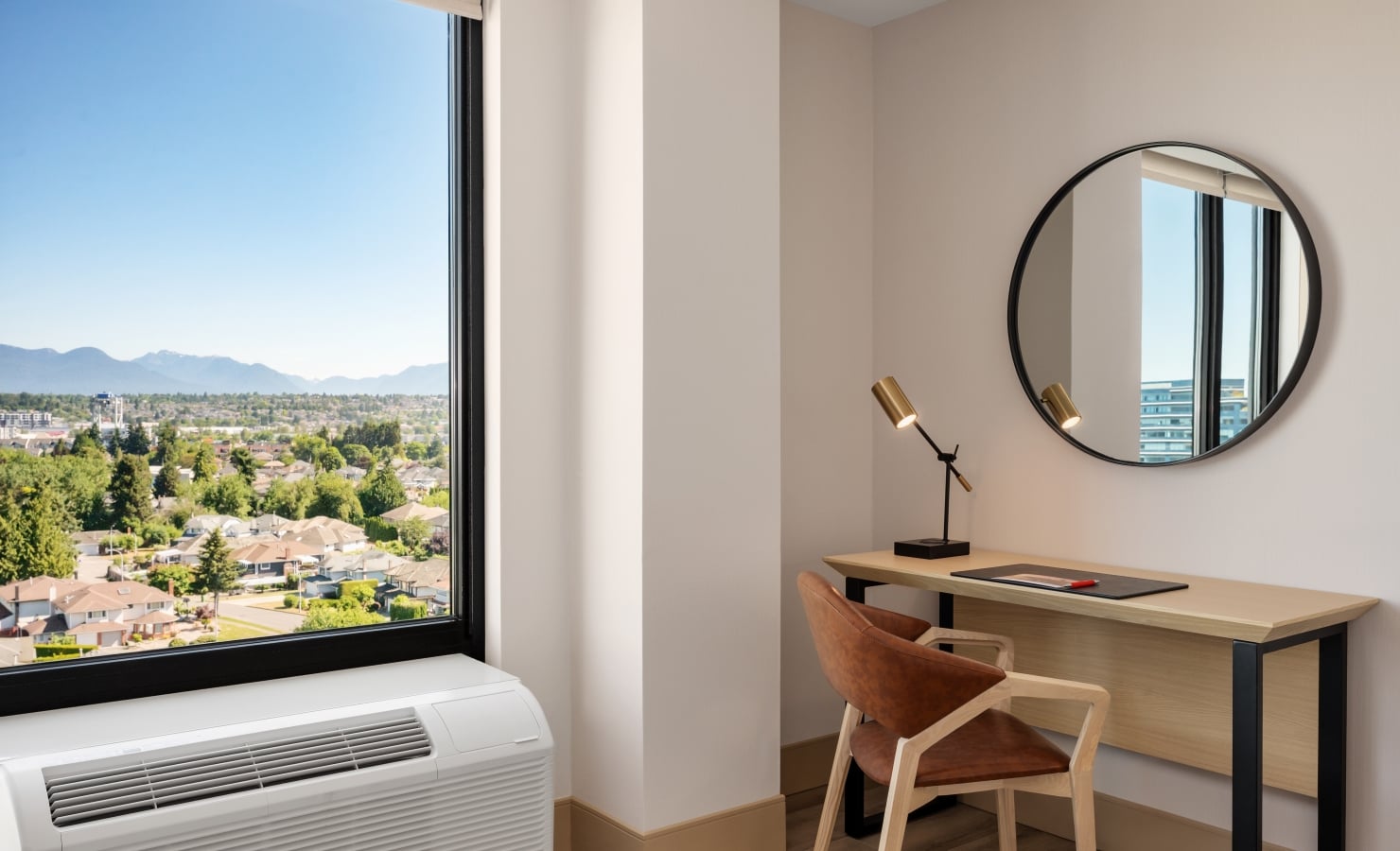 Home office corner with a wooden desk, round mirror, brass adjustable lamp, and a large window showing a city and distant mountains.