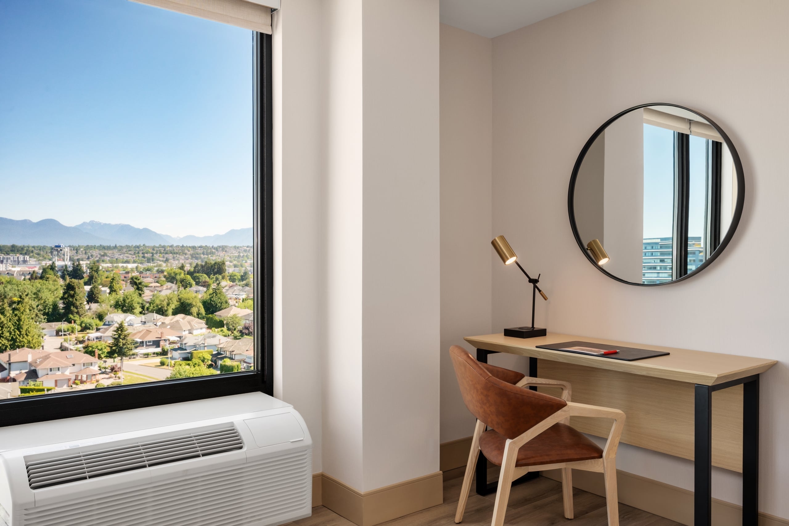 Home office corner with a wooden desk, round mirror, brass adjustable lamp, and a large window showing a city and distant mountains.