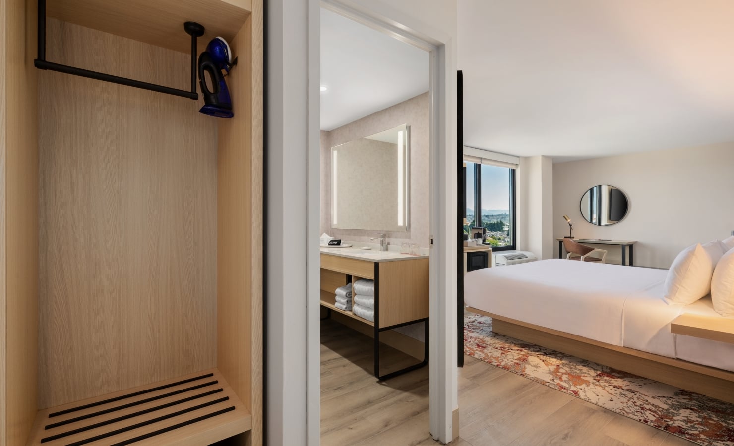 Open wardrobe area leading to a bathroom and bedroom in a hotel suite, showing a bed, rug, round mirror, and window view.
