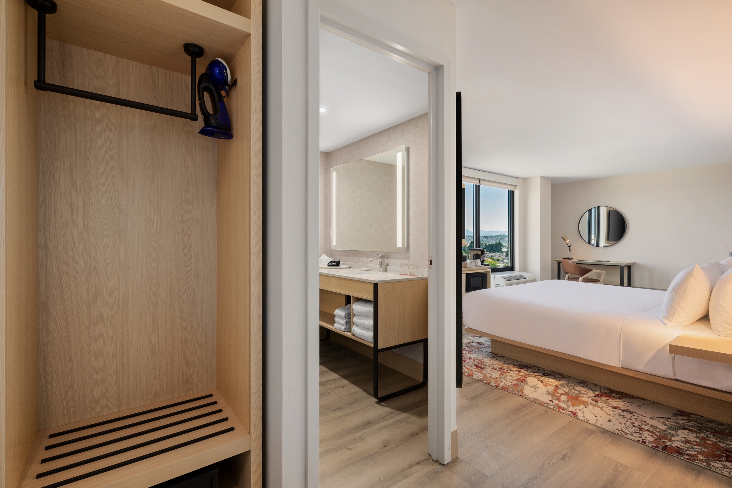 Open wardrobe area leading to a bathroom and bedroom in a hotel suite, showing a bed, rug, round mirror, and window view.