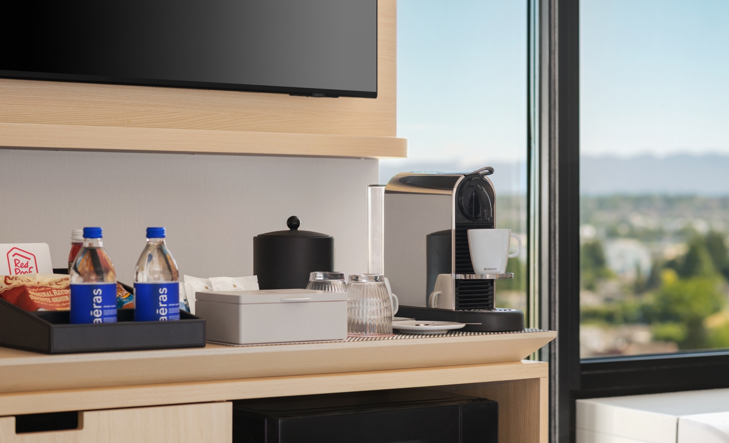 Kitchen counter with a coffee machine, kettle, mugs, and snacks set up near a large window with a scenic view