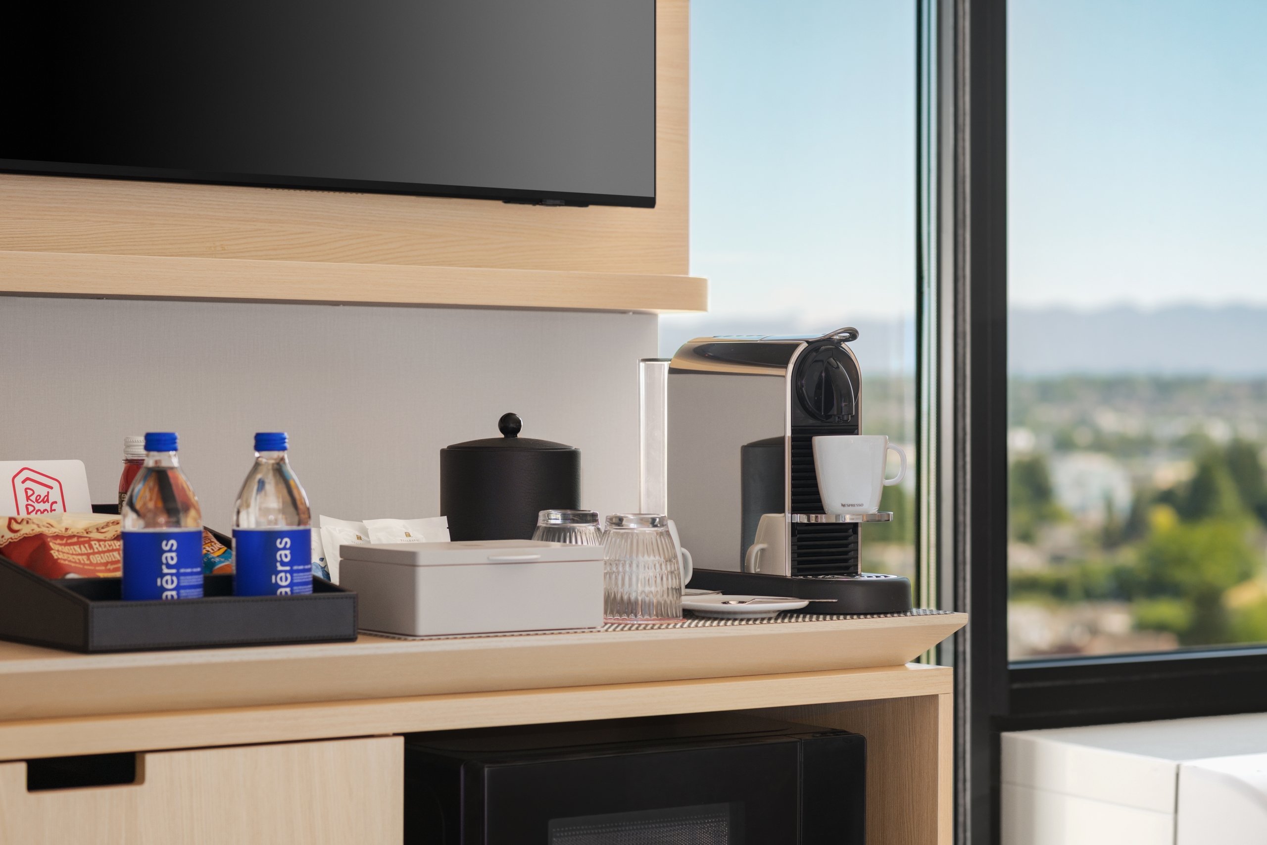 Kitchen counter with a coffee machine, kettle, mugs, and snacks set up near a large window with a scenic view