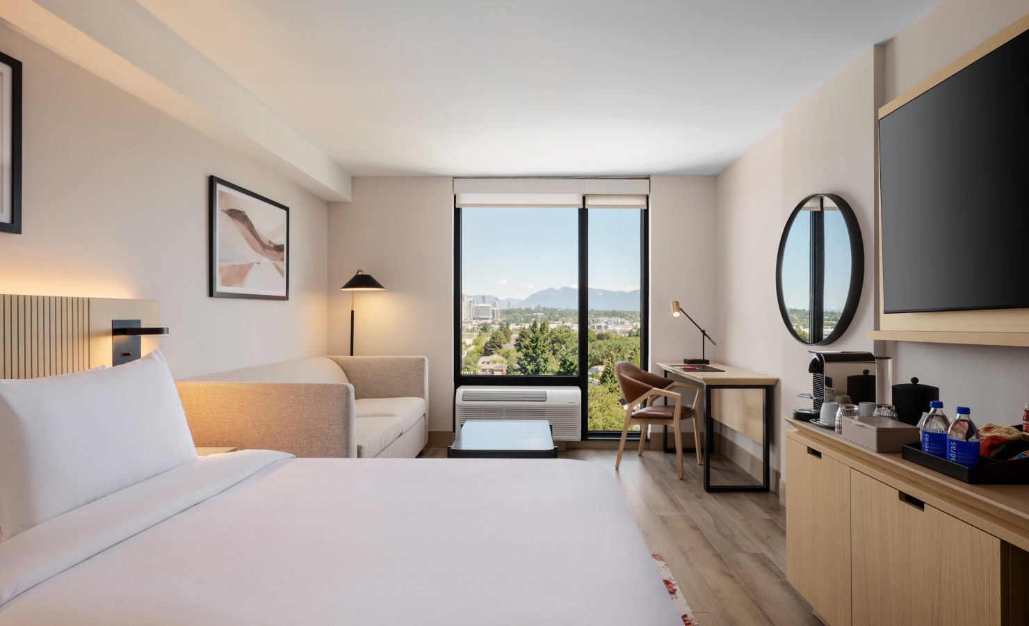 Modern hotel room with a large white bed in the foreground, beige sofa, and a desk by a window showing a city and mountain view.