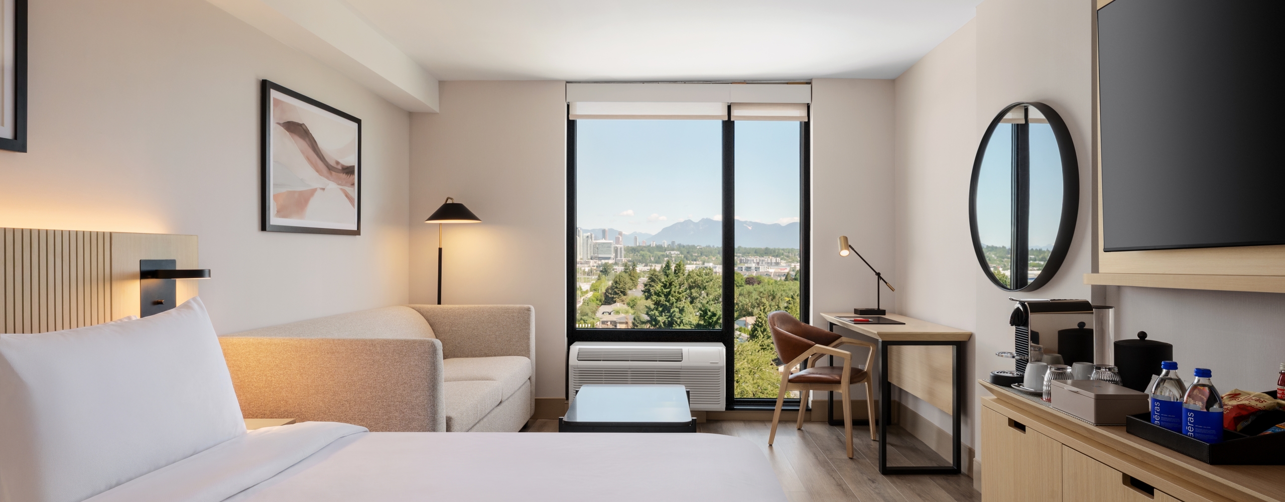 Modern hotel room with a large white bed in the foreground, beige sofa, and a desk by a window showing a city and mountain view.