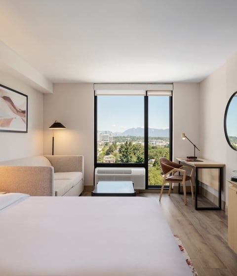 Modern hotel room with a large white bed in the foreground, beige sofa, and a desk by a window showing a city and mountain view.