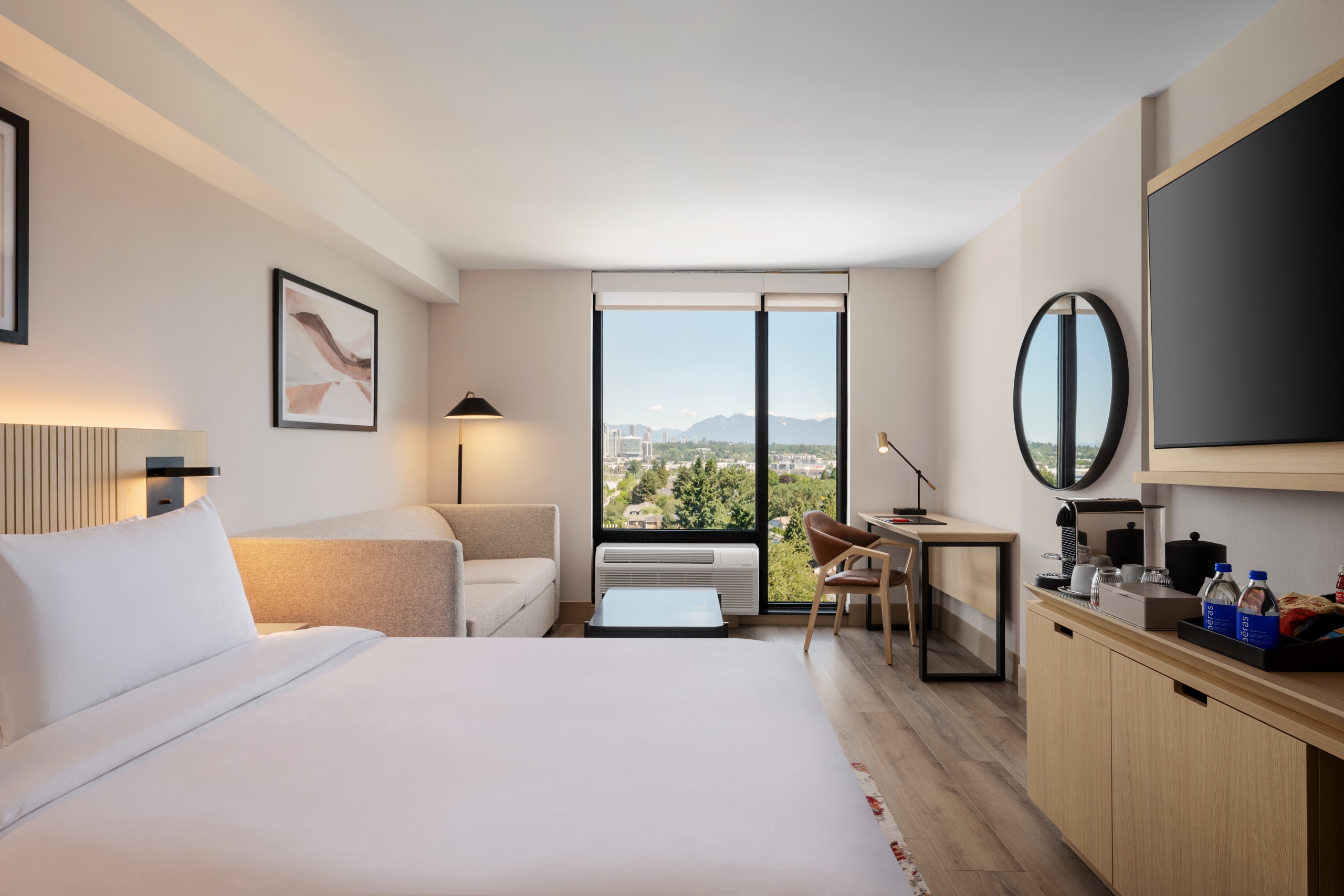 Modern hotel room with a large white bed in the foreground, beige sofa, and a desk by a window showing a city and mountain view.