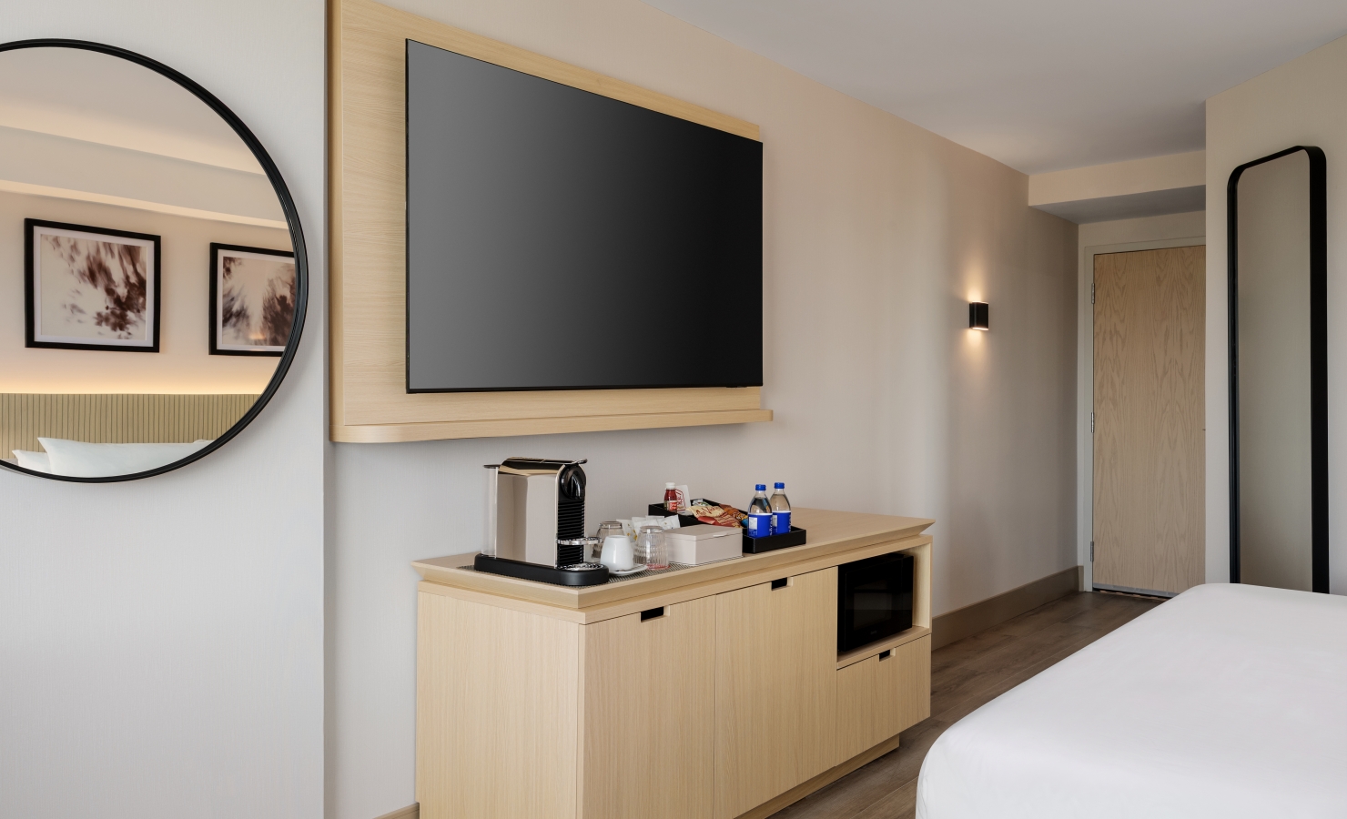 Hotel room interior with a wall-mounted flat-screen TV on a wooden panel above a dresser and a round wall mirror on the left.