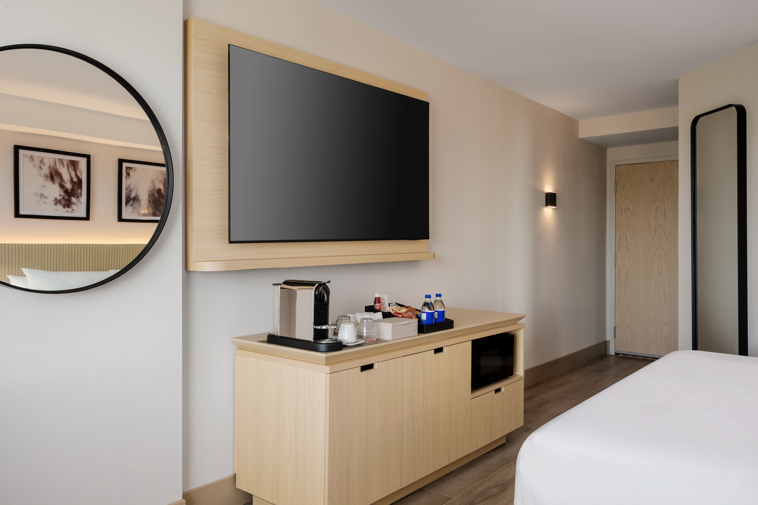 Hotel room interior with a wall-mounted flat-screen TV on a wooden panel above a dresser and a round wall mirror on the left.
