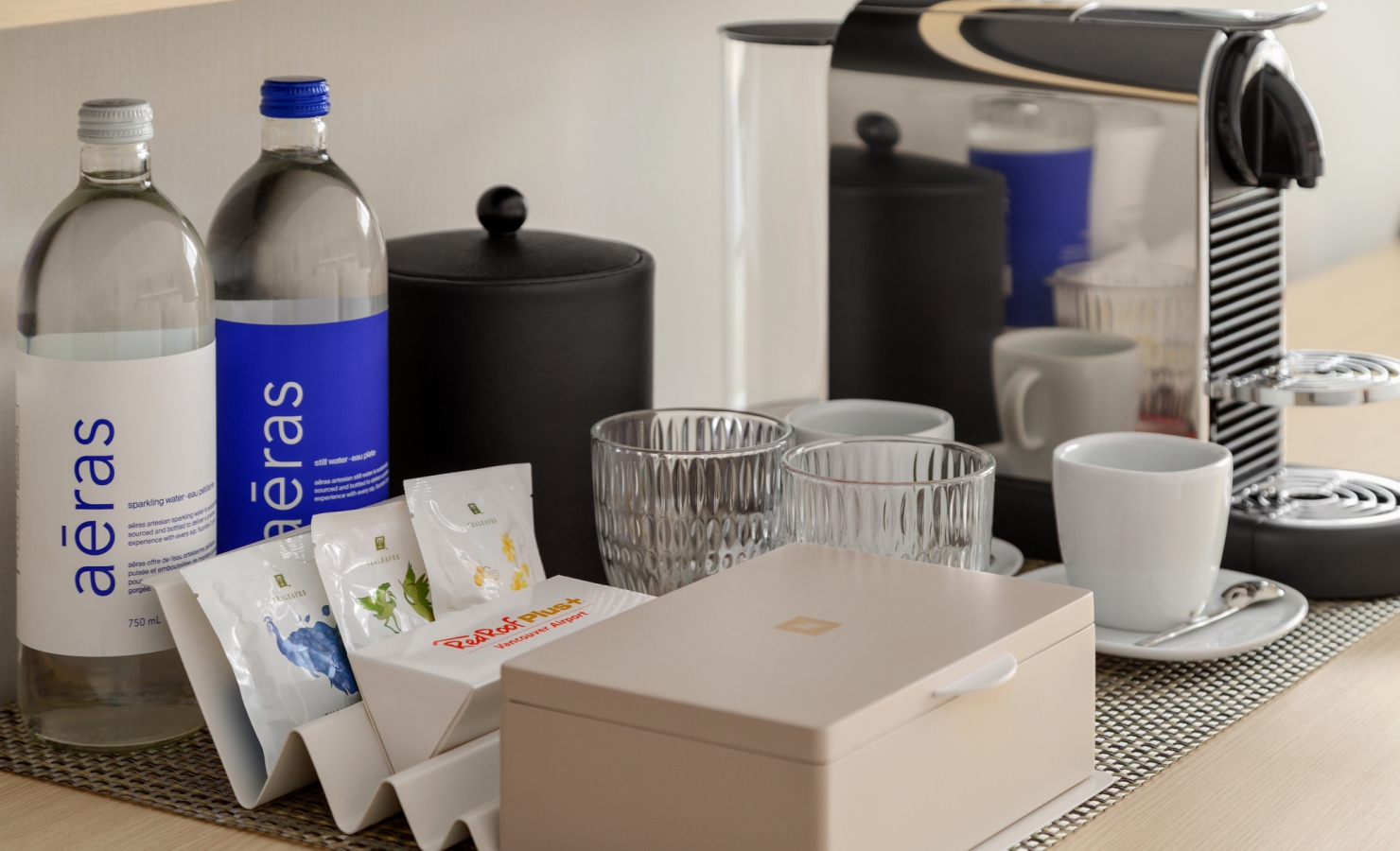 Coffee station with a silver espresso machine, two water bottles, glass cups, and assorted sachets on a woven mat.