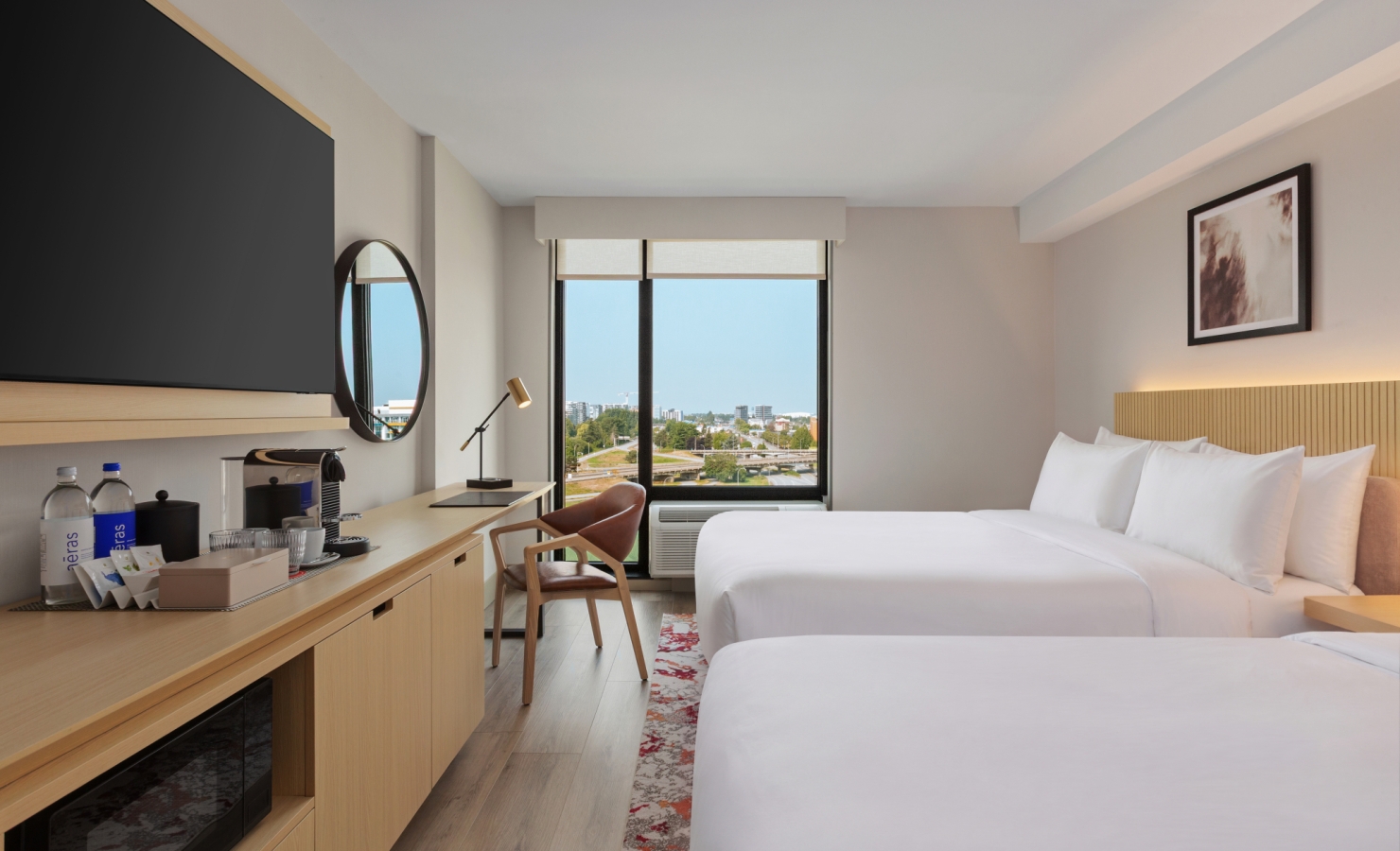 Hotel room with two white beds, wooden desk and wall-mounted TV, and a panoramic window view of the city outside the window.
