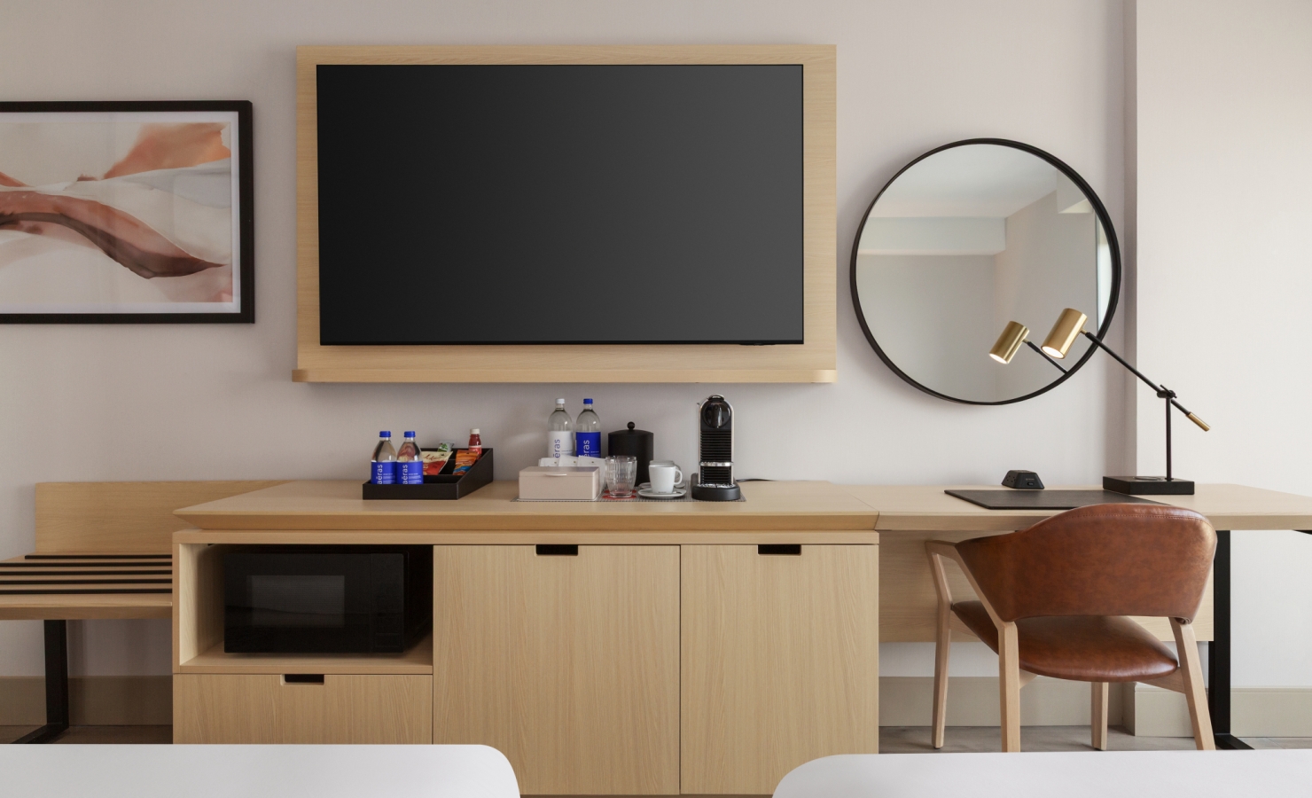 Wall-mounted flat-screen TV above a wooden cabinet with a coffee setup, round mirror and desk lamp on the right.