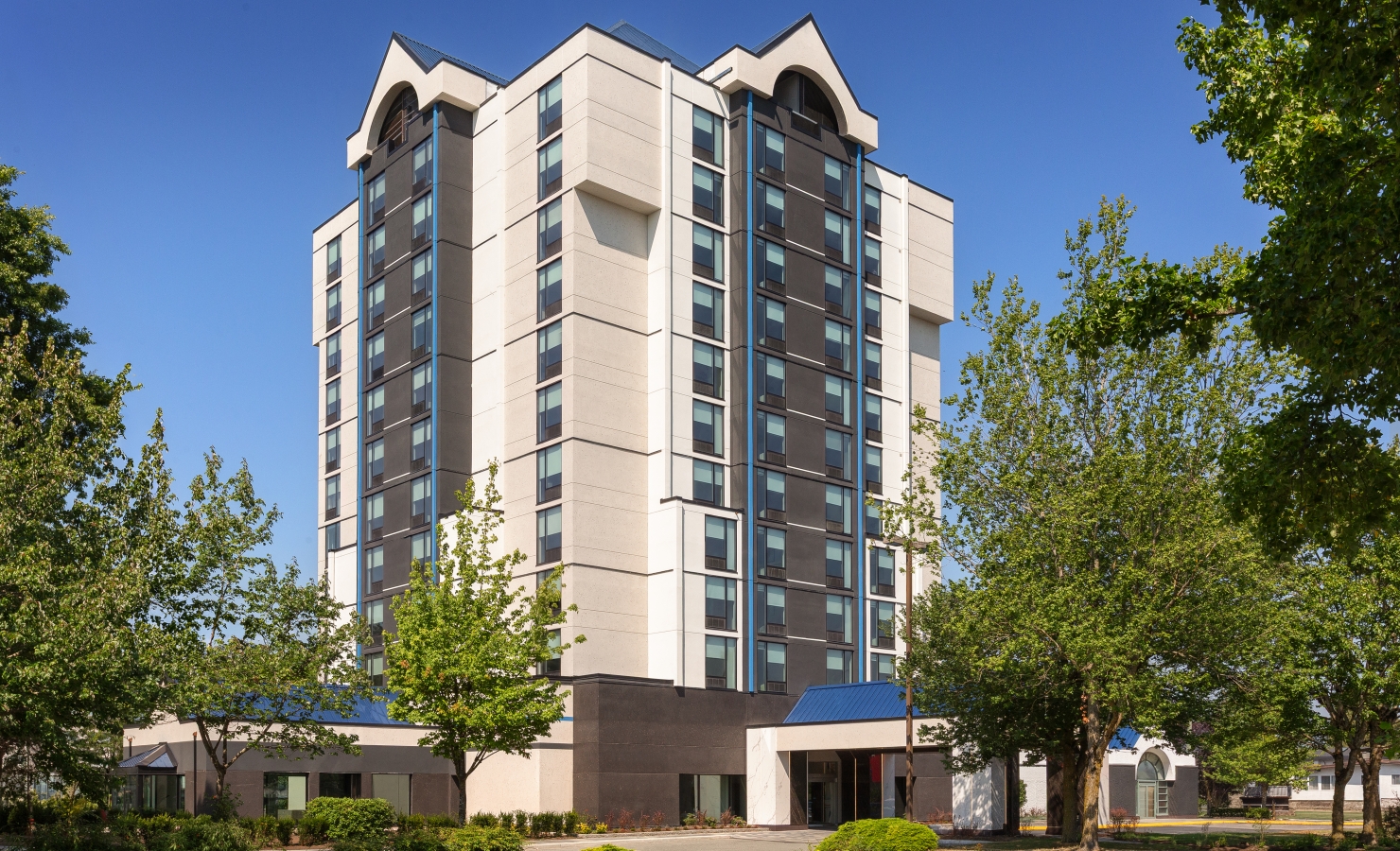 Modern high-rise apartment building with tan and dark gray panels, glass windows, and surrounding green trees against a blue sky.