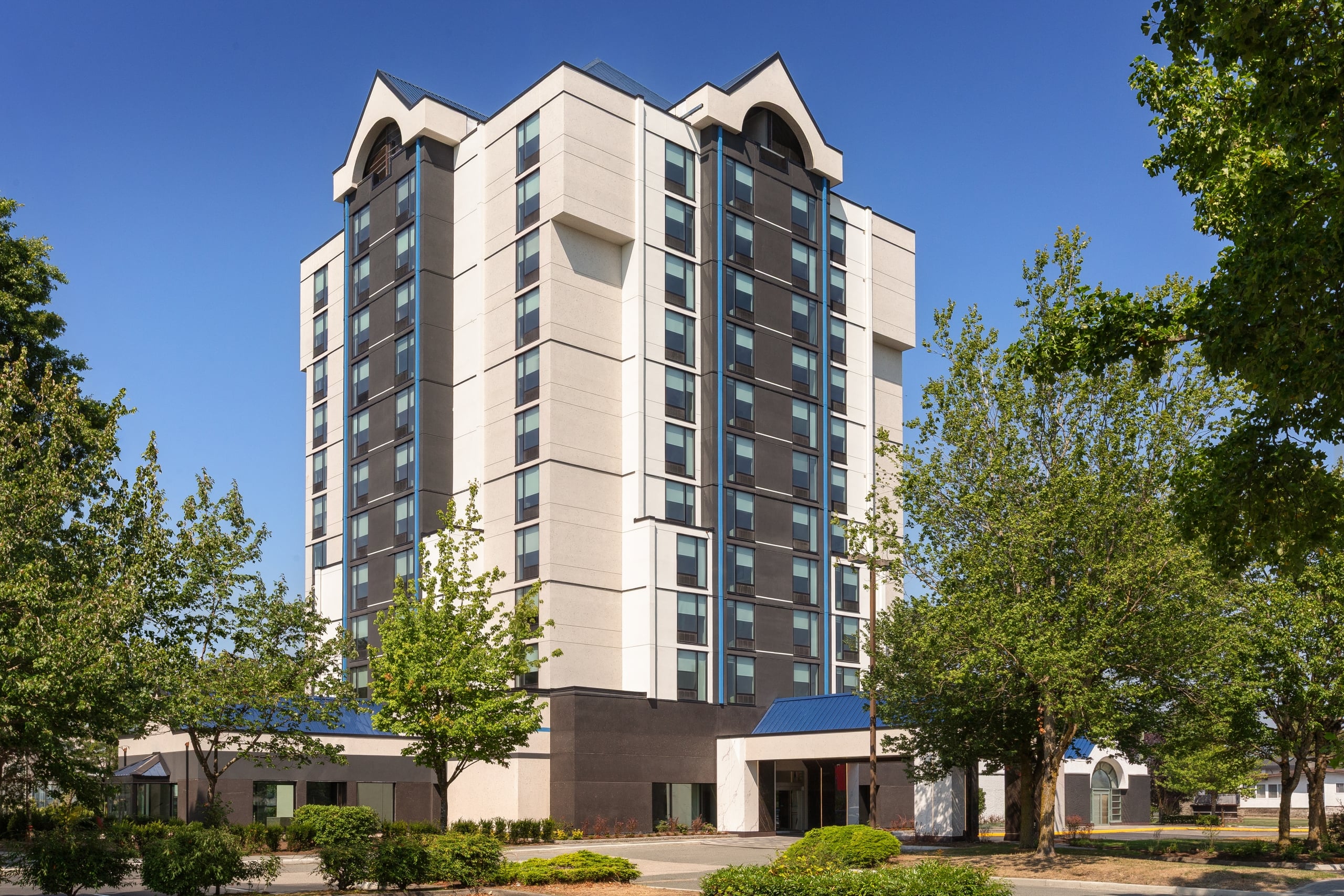 Modern high-rise apartment building with tan and dark gray panels, glass windows, and surrounding green trees against a blue sky.
