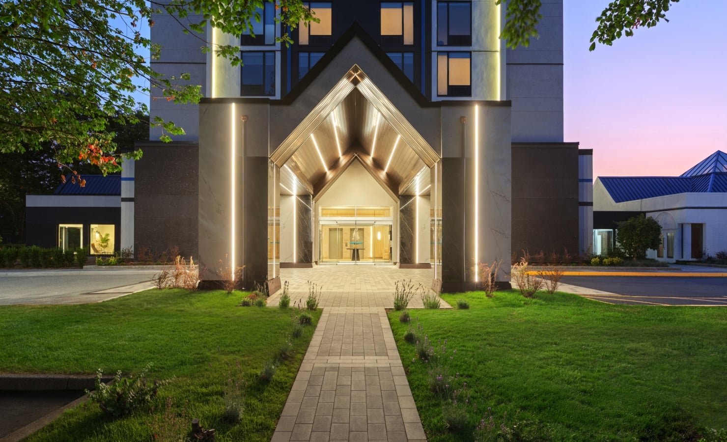 Modern building entrance with a bright glass triangular canopy and warm interior lights, framed by tall stone columns at dusk.