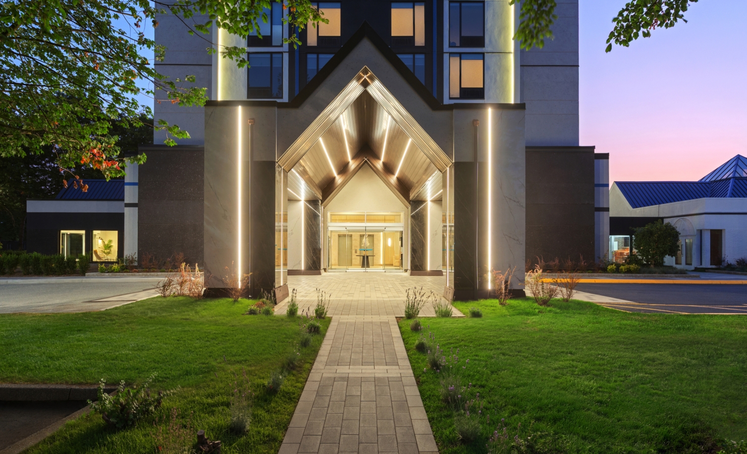 Modern building entrance with a glowing triangular canopy, glass doors, and warm interior lighting, framed by stone columns on a green lawn.