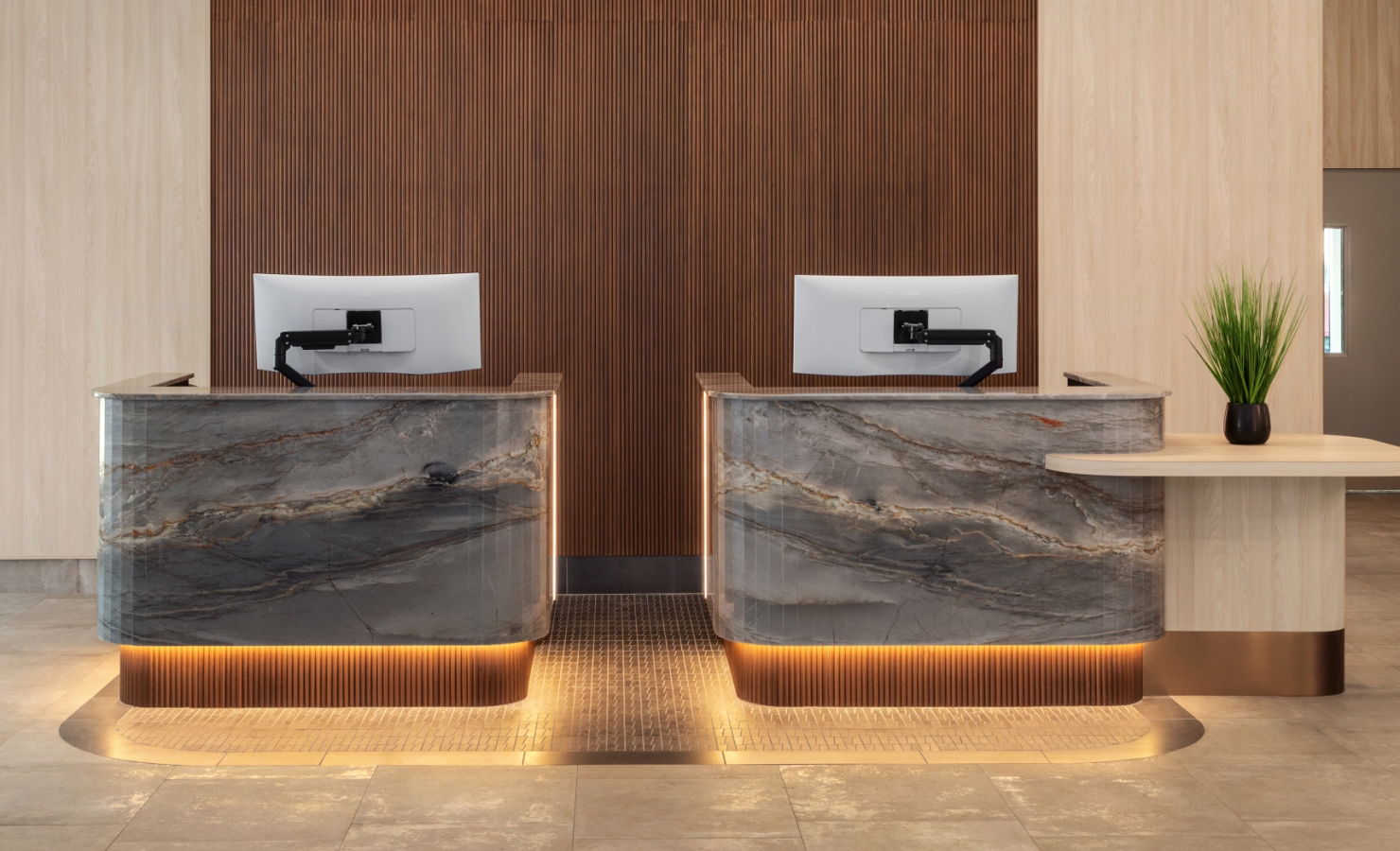 Reception desk with two marble counters in a wood-paneled backdrop, computer monitors, and a potted plant on the right, warm ambient lighting.