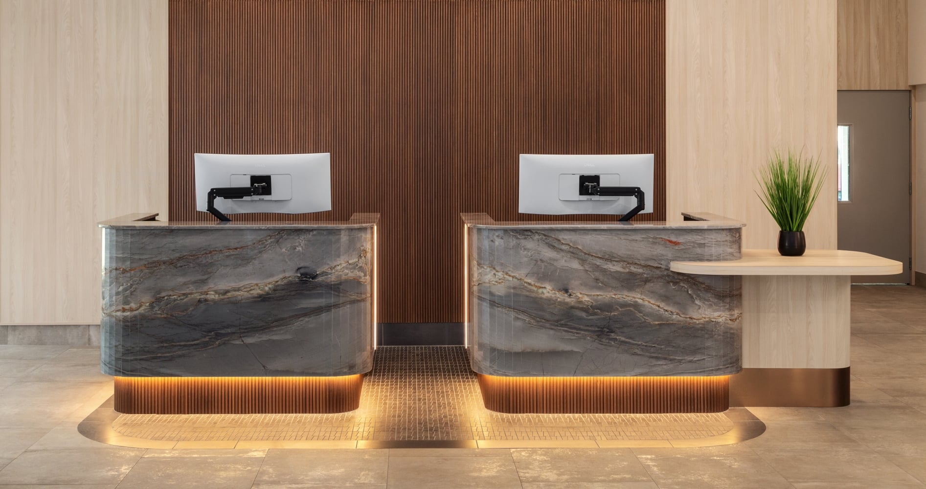 Reception desk with two marble counters in a wood-paneled backdrop, computer monitors, and a potted plant on the right, warm ambient lighting.
