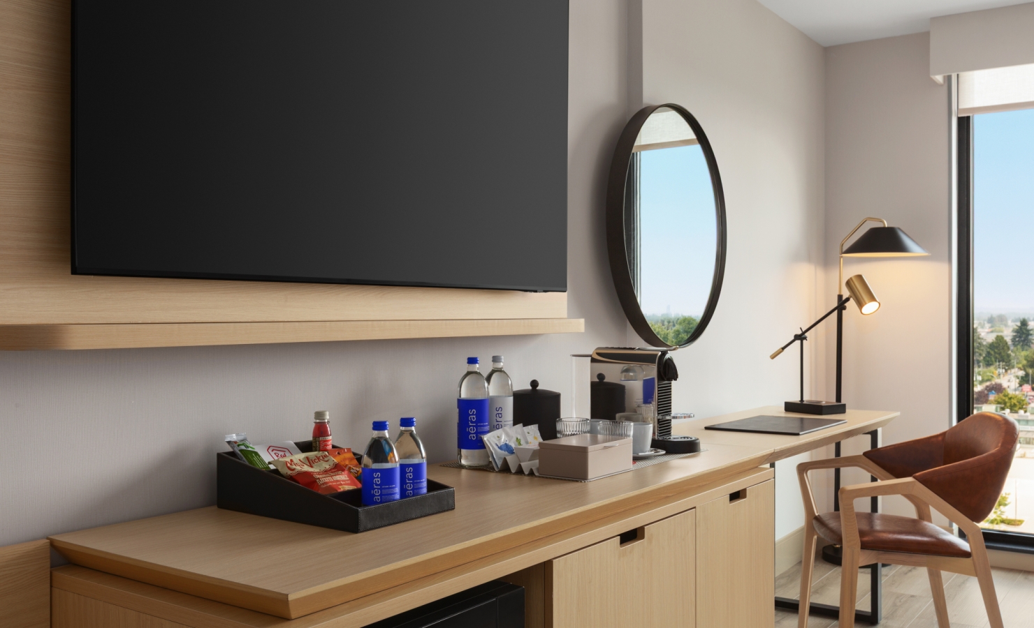 Hotel kitchenette area with a mounted flat-screen TV, round wall mirror, and a snack/drink station on a wooden counter.