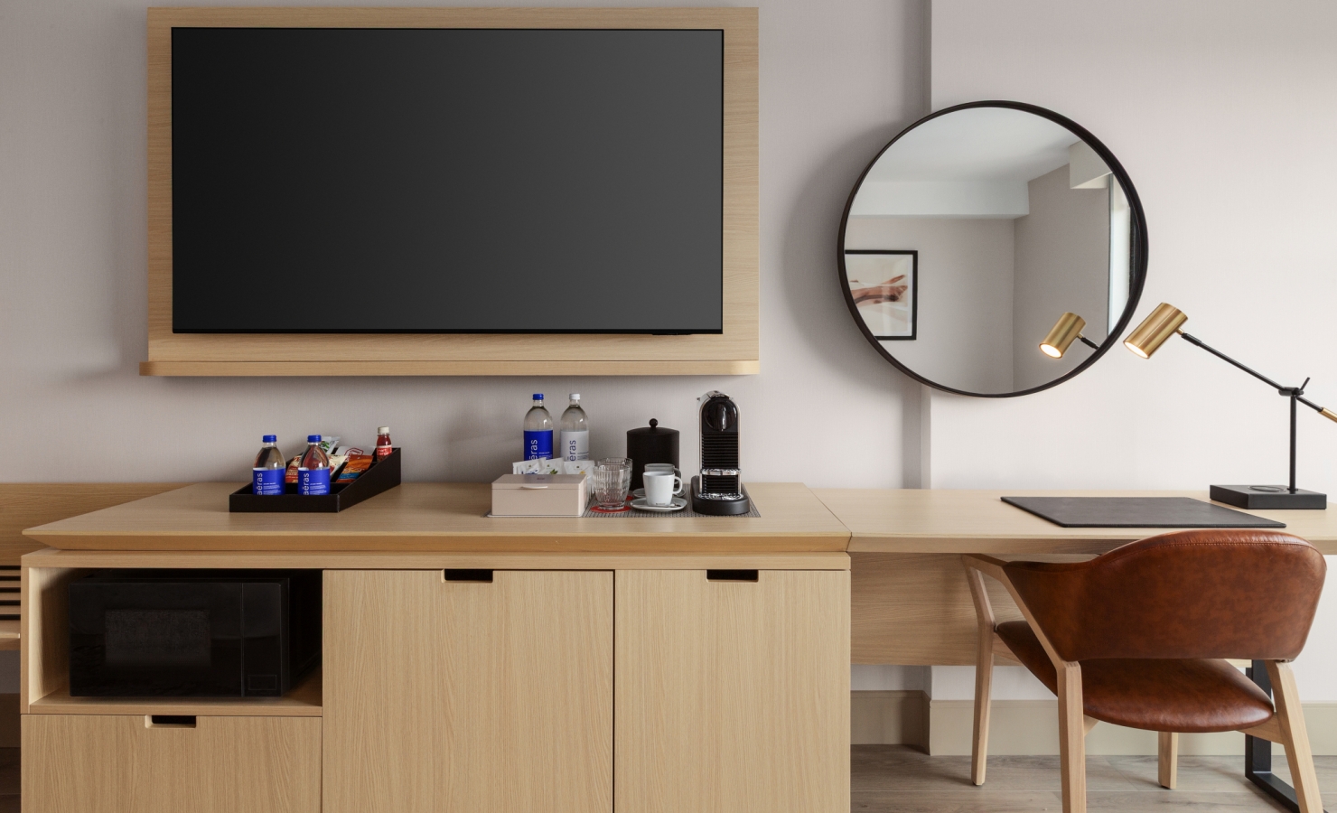 Hotel minibar and coffee station on a wooden cabinet beneath a wall-mounted flat-screen TV; round mirror to the right.