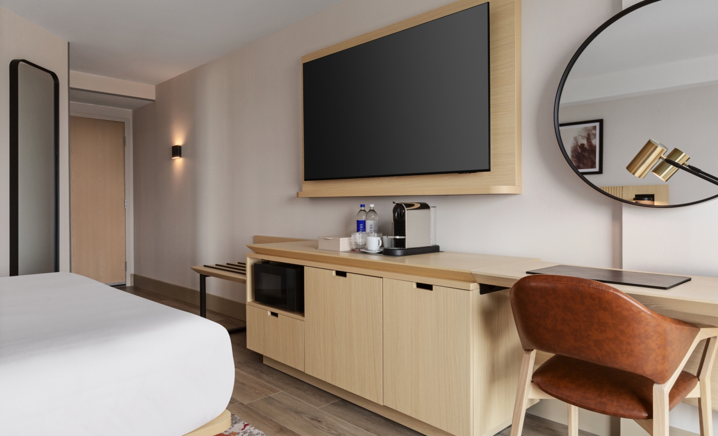 Hotel room interior with a large flat-screen TV in a light wood frame above a wooden cabinet, coffee station, and a round mirror.