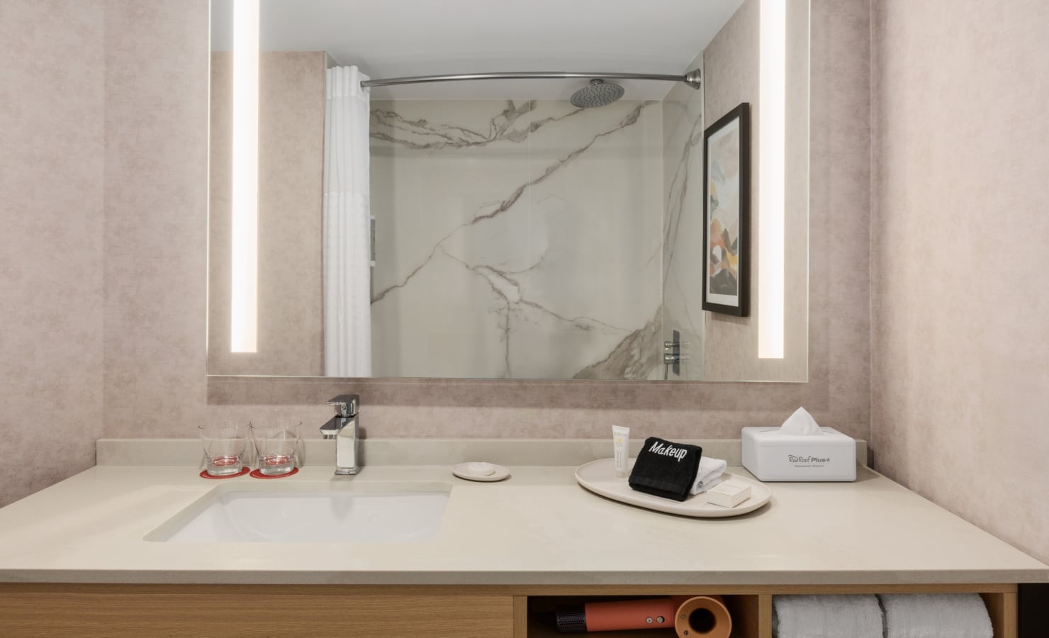 Modern bathroom vanity with a white countertop, sink, and chrome faucet, mirrored by a large wall mirror with vertical lights on each side; marble-tiled shower in the background and towels stored in an open shelf beneath.