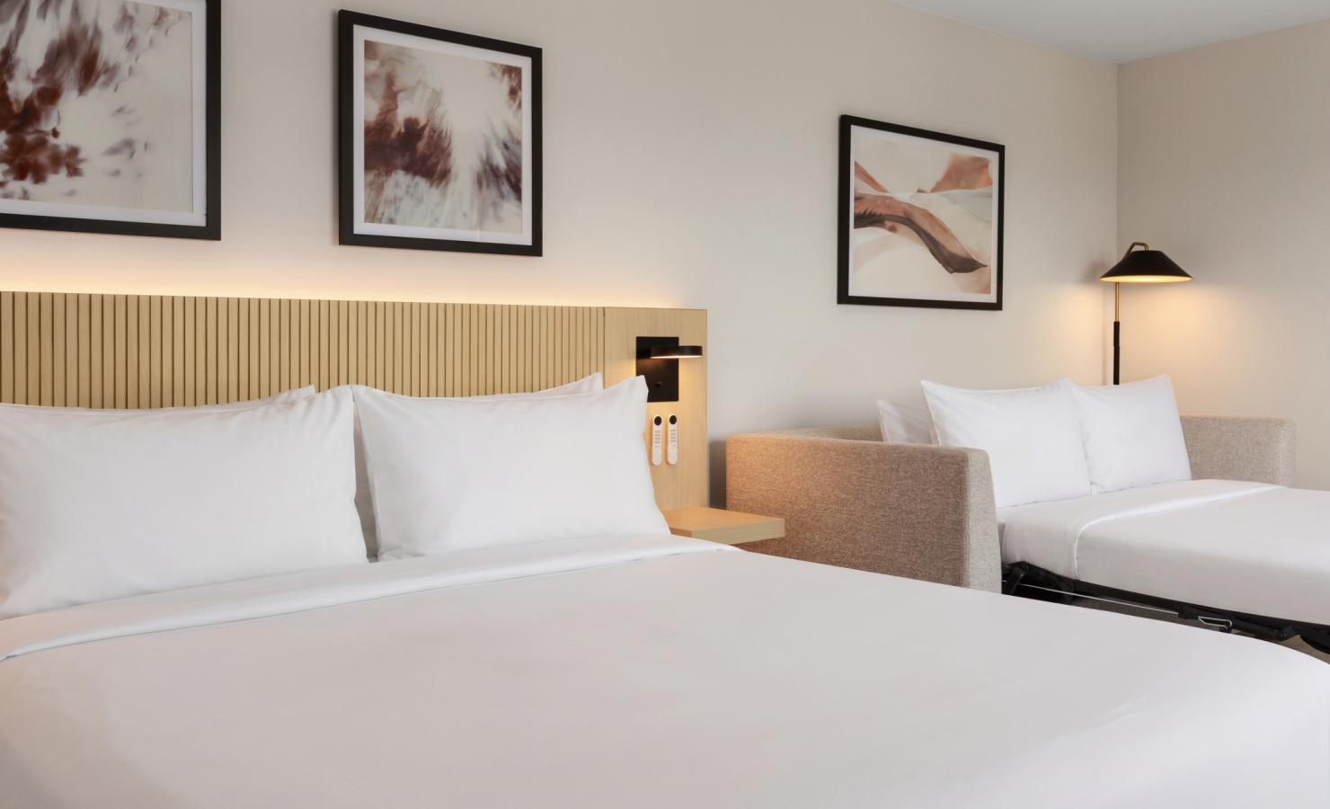 Hotel room with two white beds, beige headboard, and framed abstract art on the wall behind them.