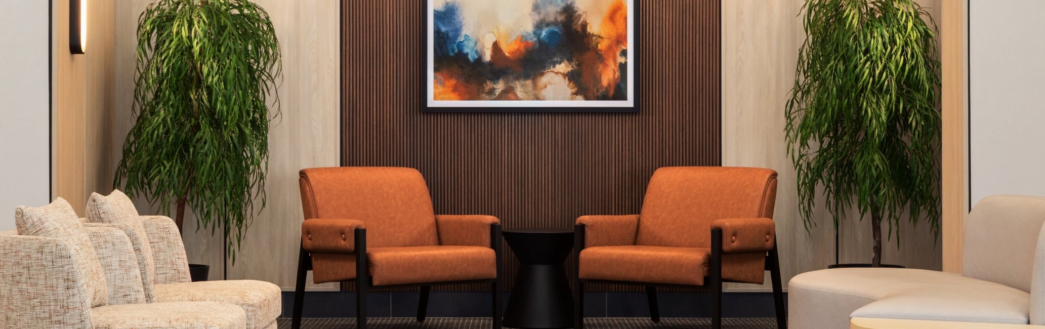 Modern hotel-style lounge with a wood-paneled accent wall, abstract framed painting, orange chairs, and green potted plants.